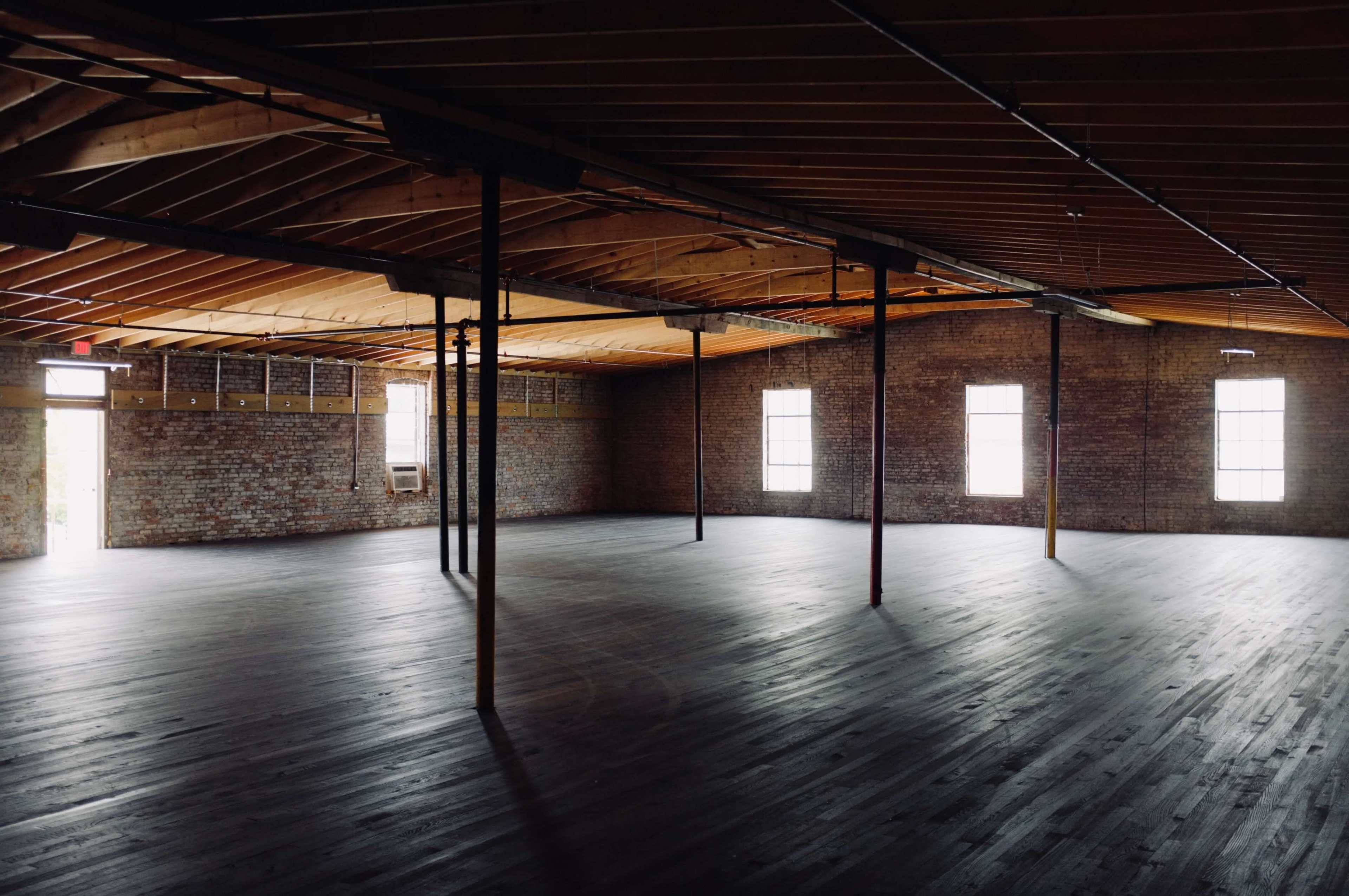 An empty, industrial-style room features exposed brick walls, wooden beams overhead, and large windows allowing natural light to enter.