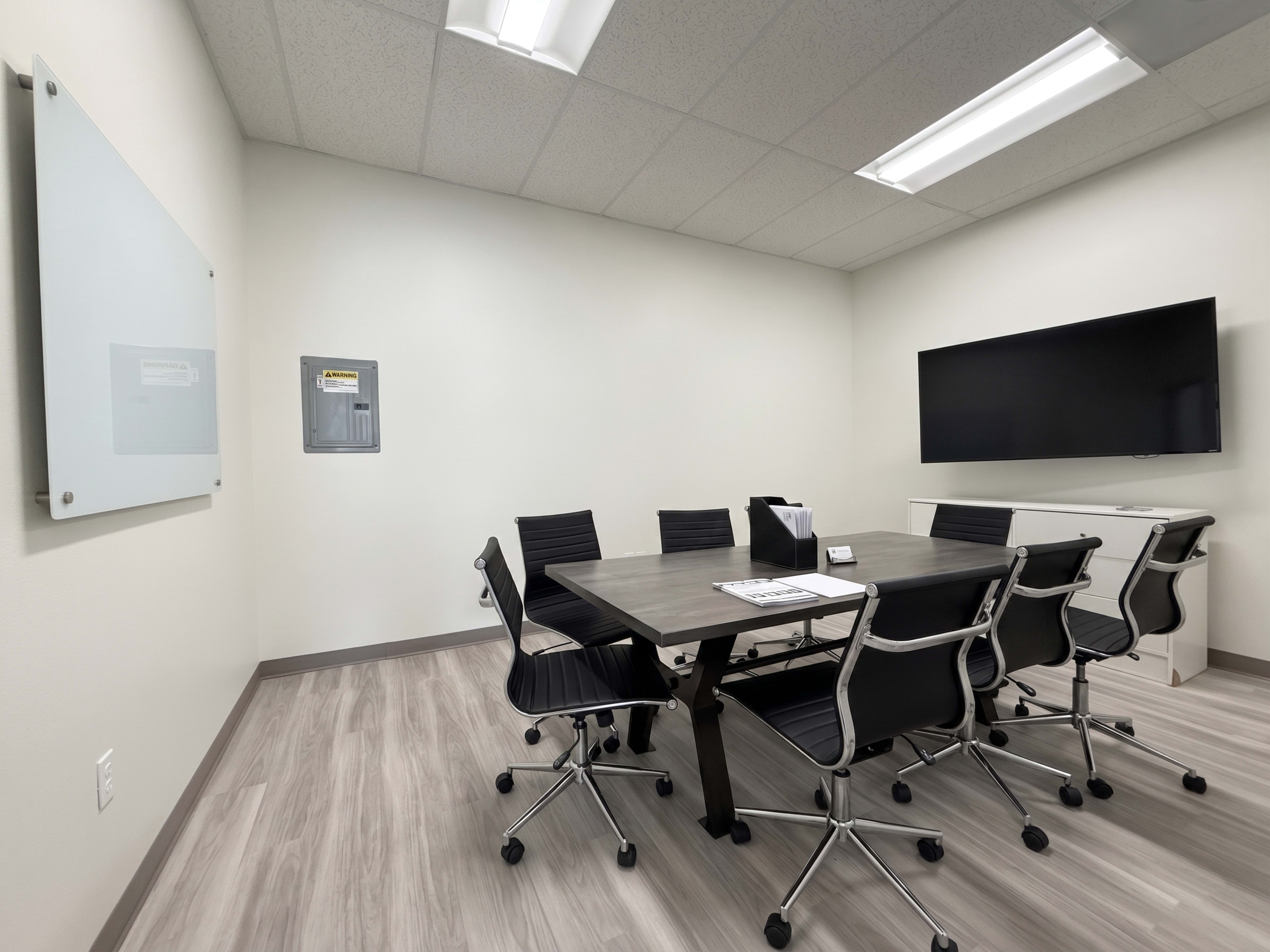 A modern conference room features a rectangular table surrounded by black ergonomic chairs, a wall-mounted television, and a whiteboard.