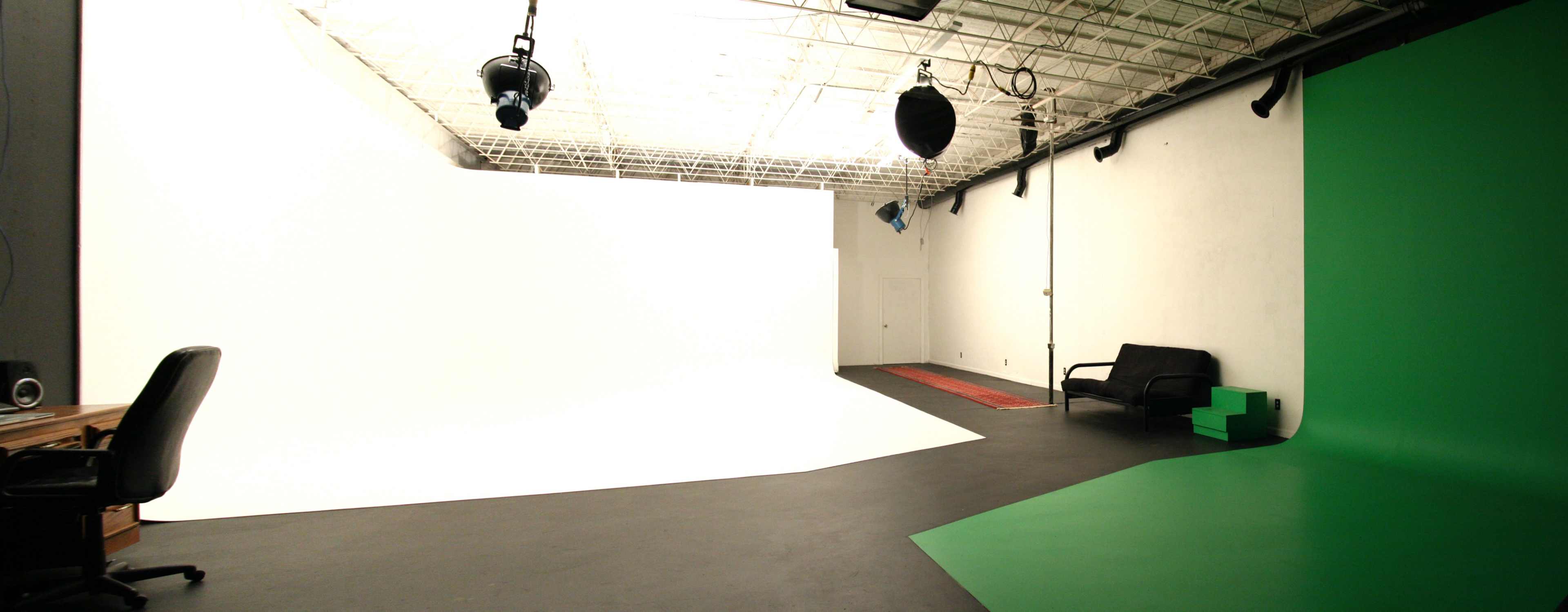 The image shows a photography studio with a white backdrop, a green backdrop, a black sofa, and a desk with a chair.