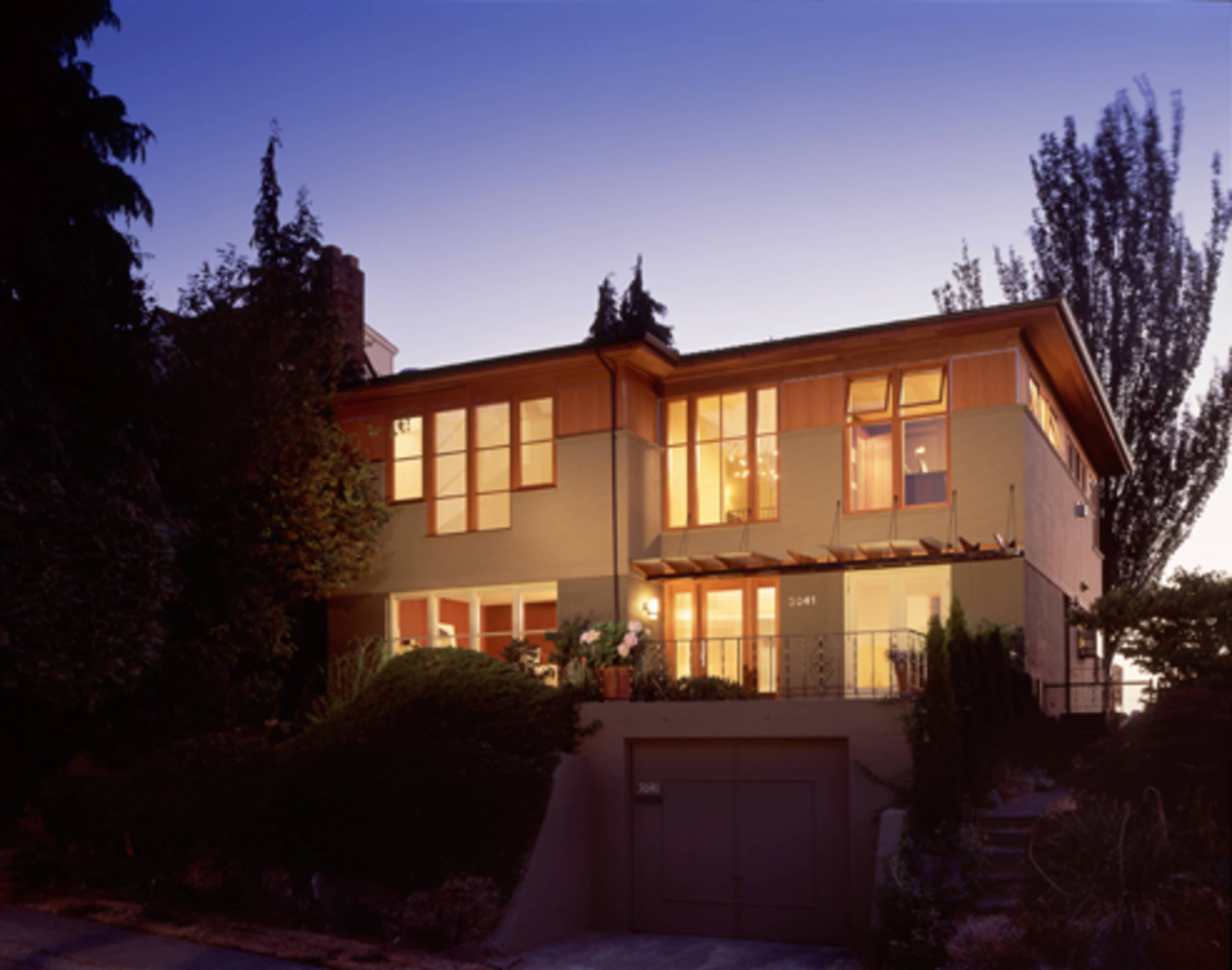 A modern two-story house with large windows and a landscaped front garden is illuminated at dusk.