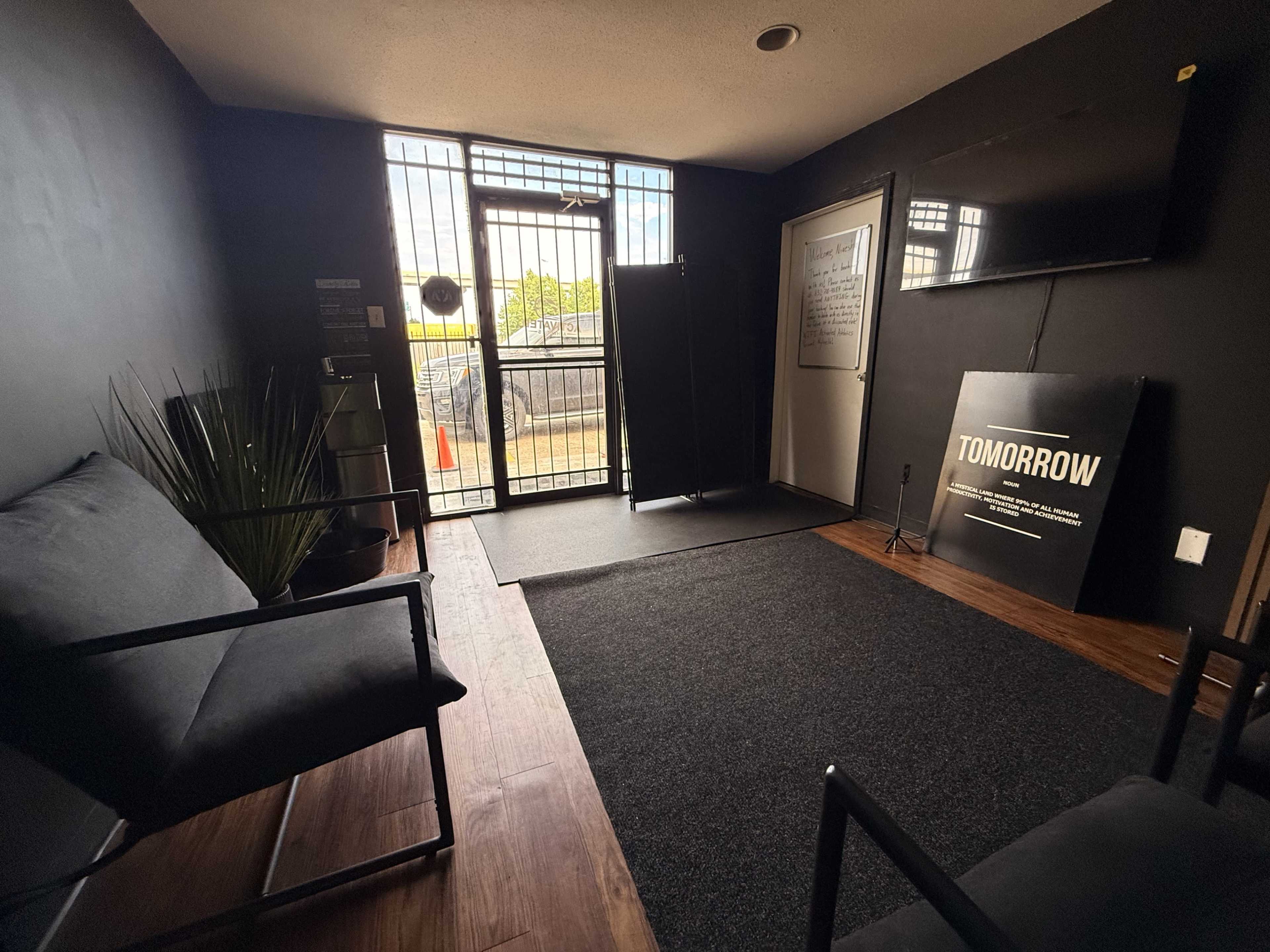 The image shows a small indoor waiting area with two black chairs, a dark rug, and a door leading outside, featuring a sign that reads "TOMORROW."