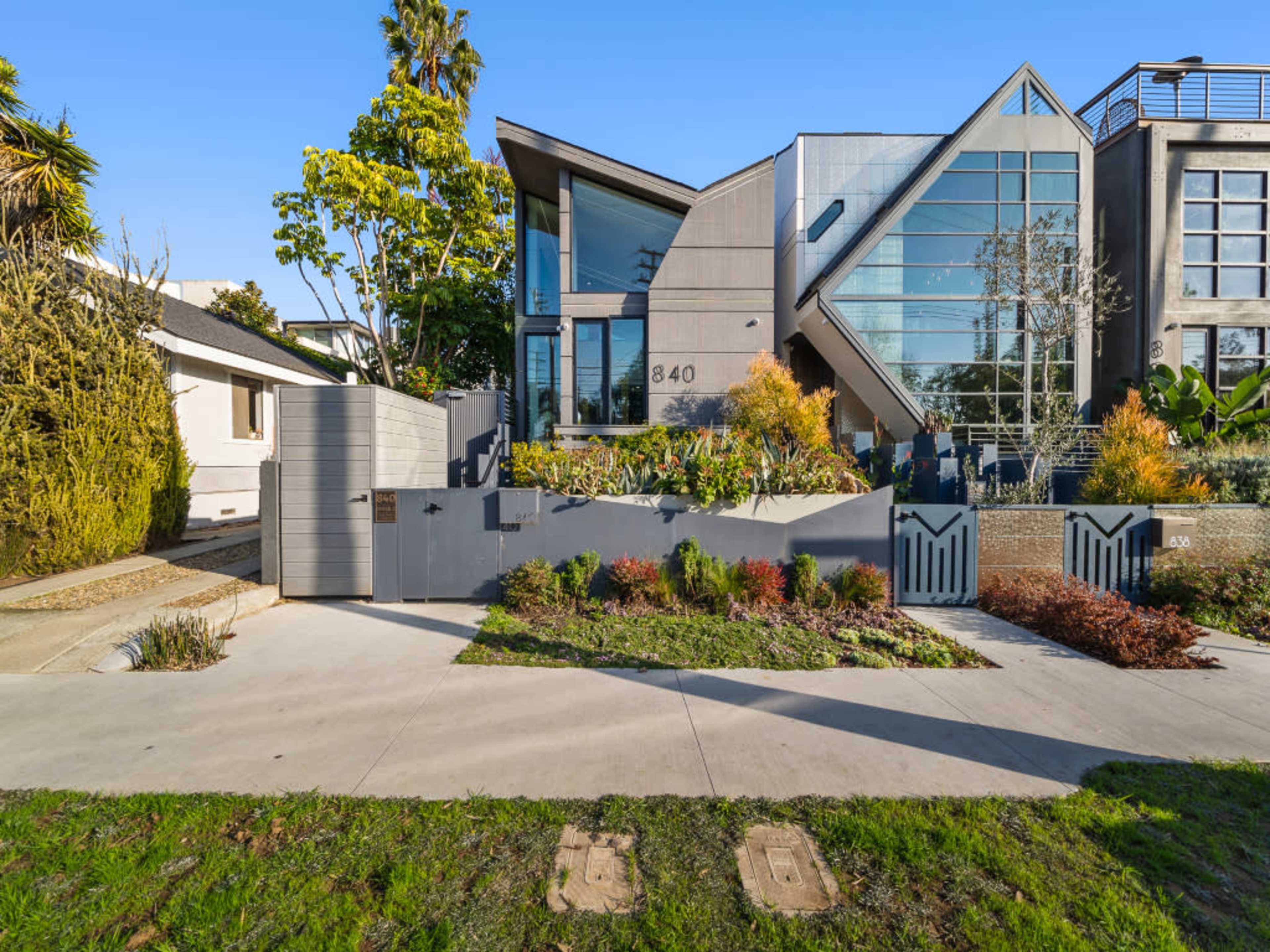 The image shows a modern house with a unique angular design, featuring large glass windows and a landscaped front yard.