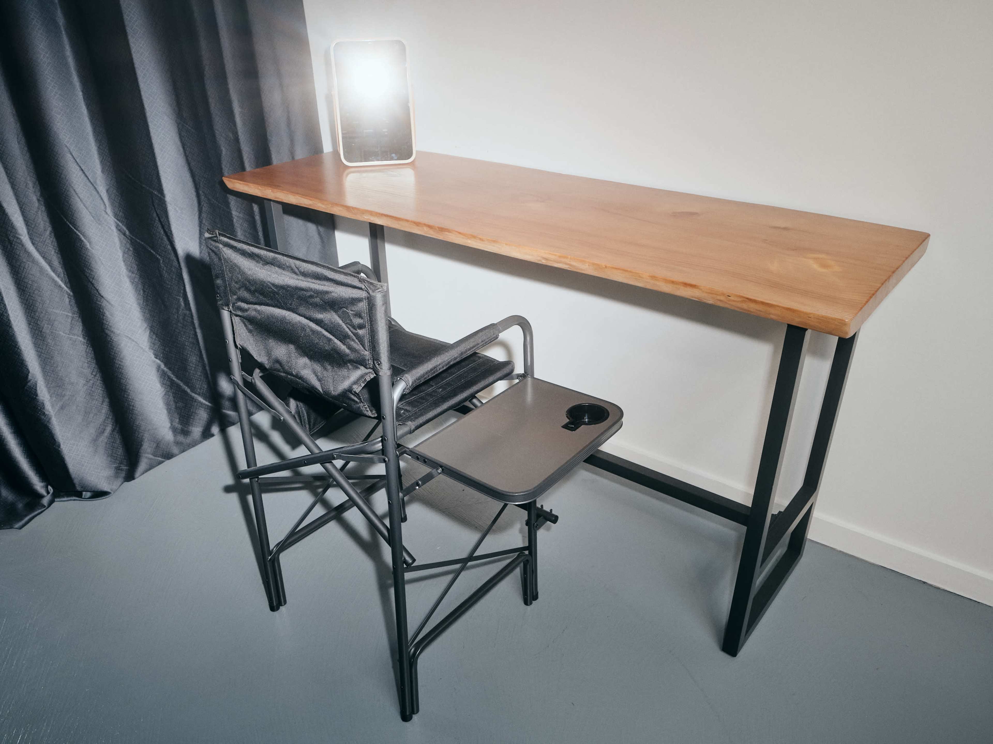 A wooden desk with a metal frame is positioned against a wall, accompanied by a folding chair and a mirror that reflects light.