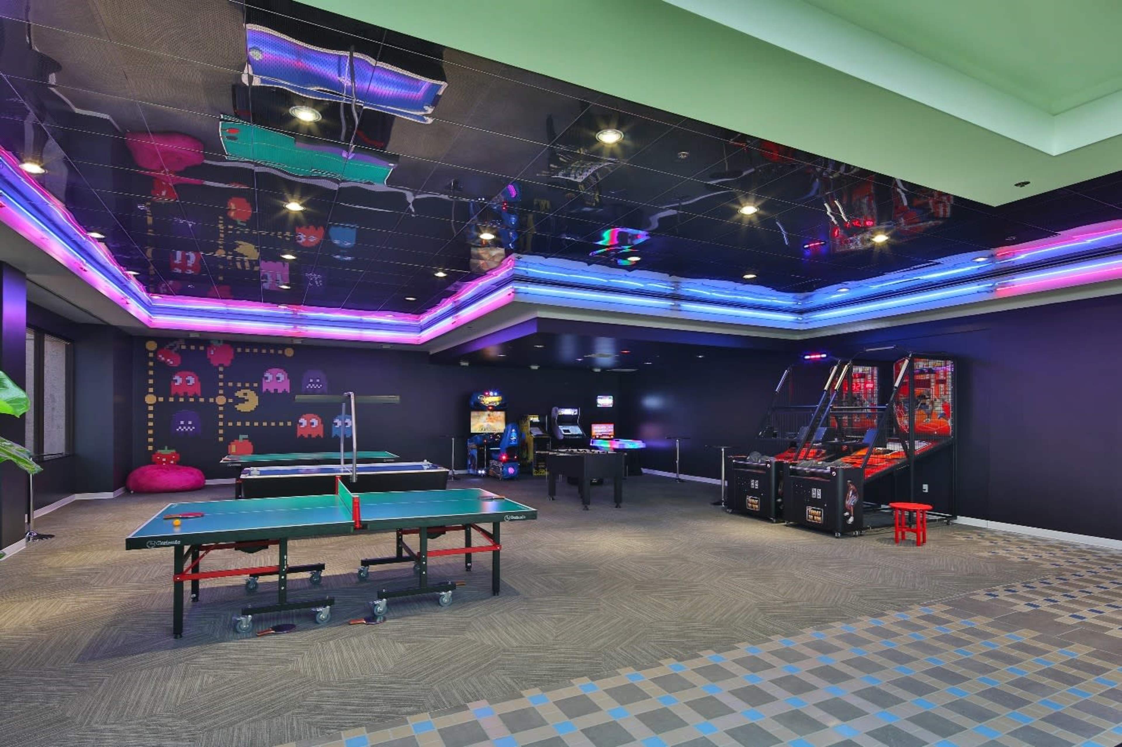 A game room featuring a ping pong table, arcade machines, and vibrant neon lights on the ceiling.