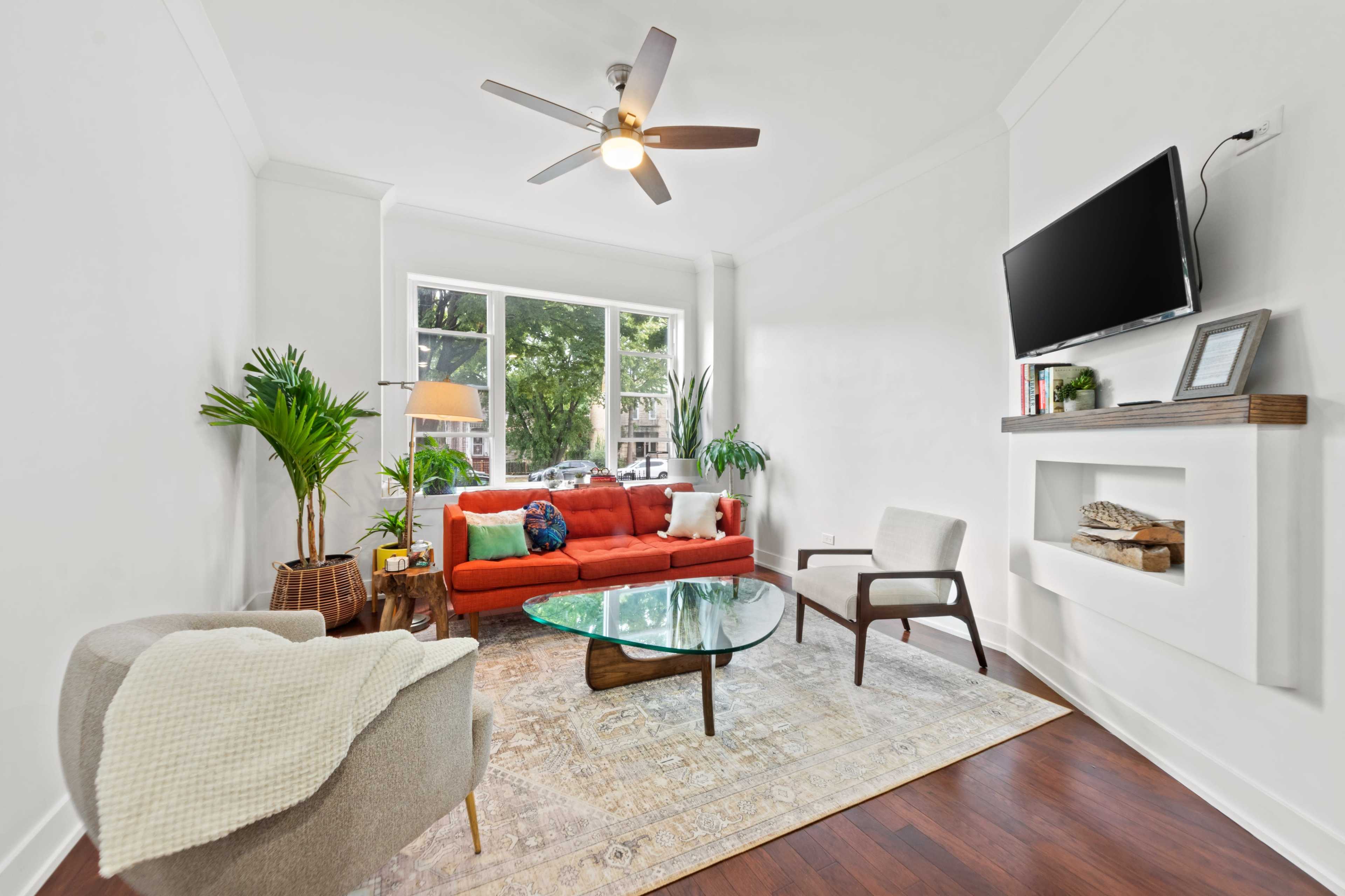 A bright living room features a red sofa, a glass coffee table, and a wall-mounted television, with large windows allowing natural light to fill the space.