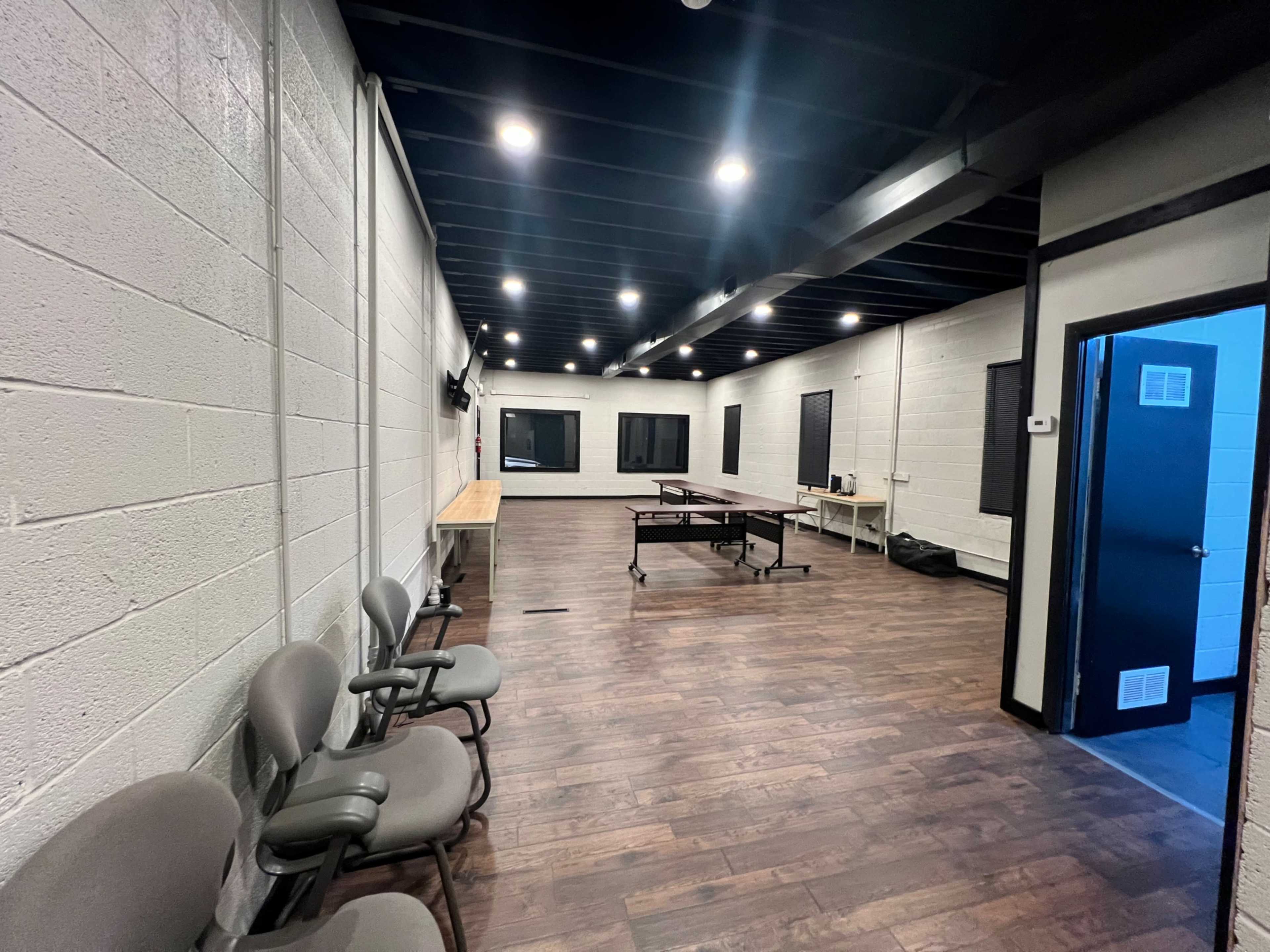 A spacious, well-lit room with wooden flooring, several chairs along one side, a long table in the center, and windows on the walls.