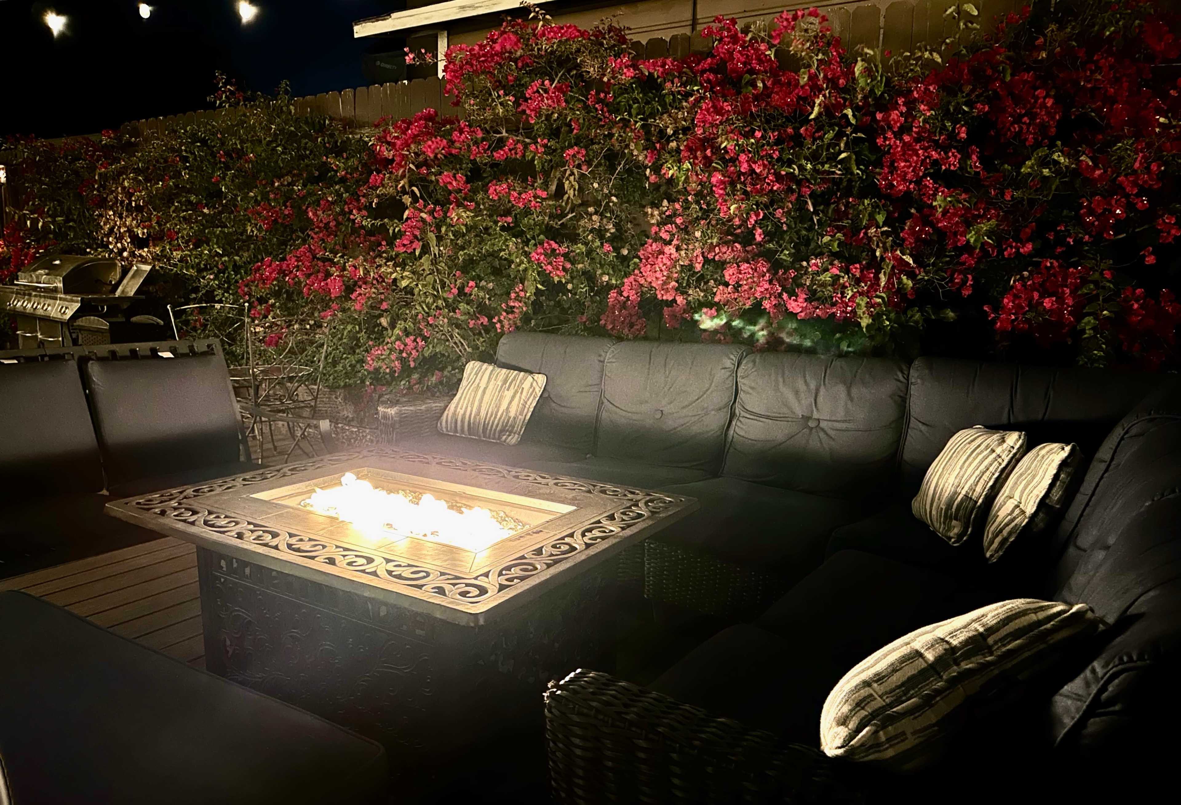 A seating area with black cushions surrounds a fire table, illuminated by nearby string lights and bordered by blooming bougainvillea.