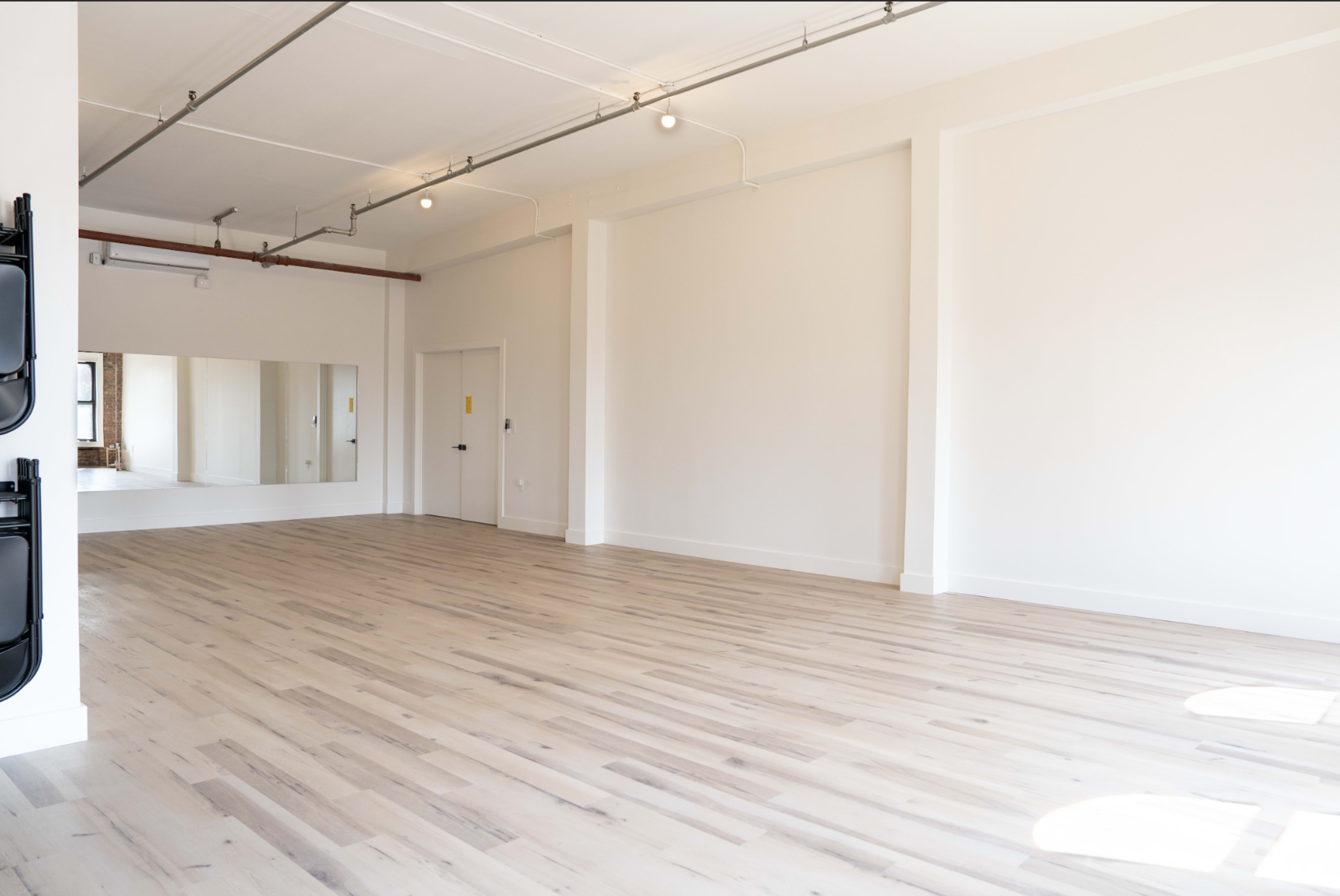The image shows a spacious, empty room with light-colored wooden flooring and a large mirror on one wall.