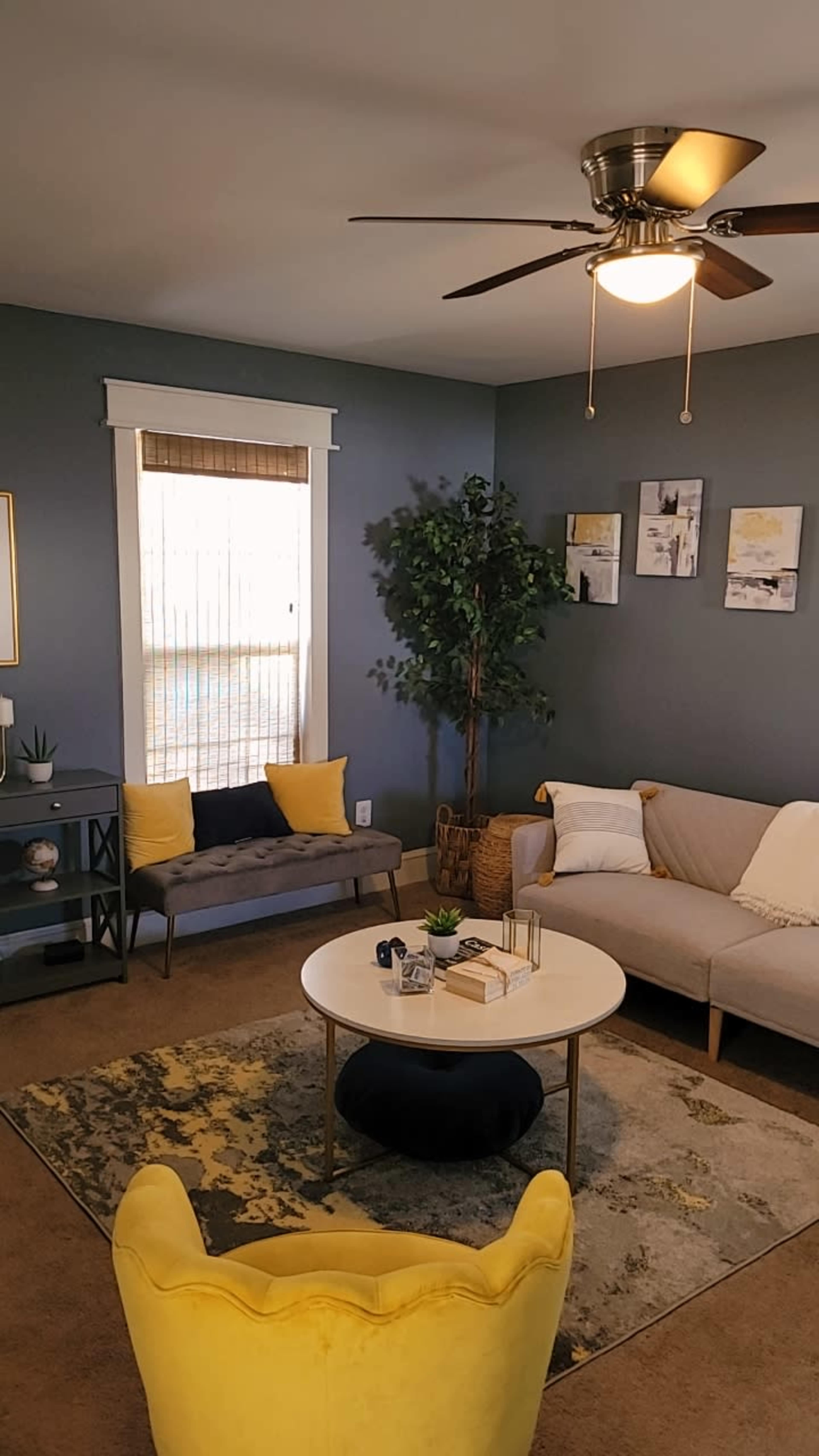 The image shows a cozy living room featuring a gray wall, a round coffee table, a yellow chair, and a small sofa with decorative pillows.