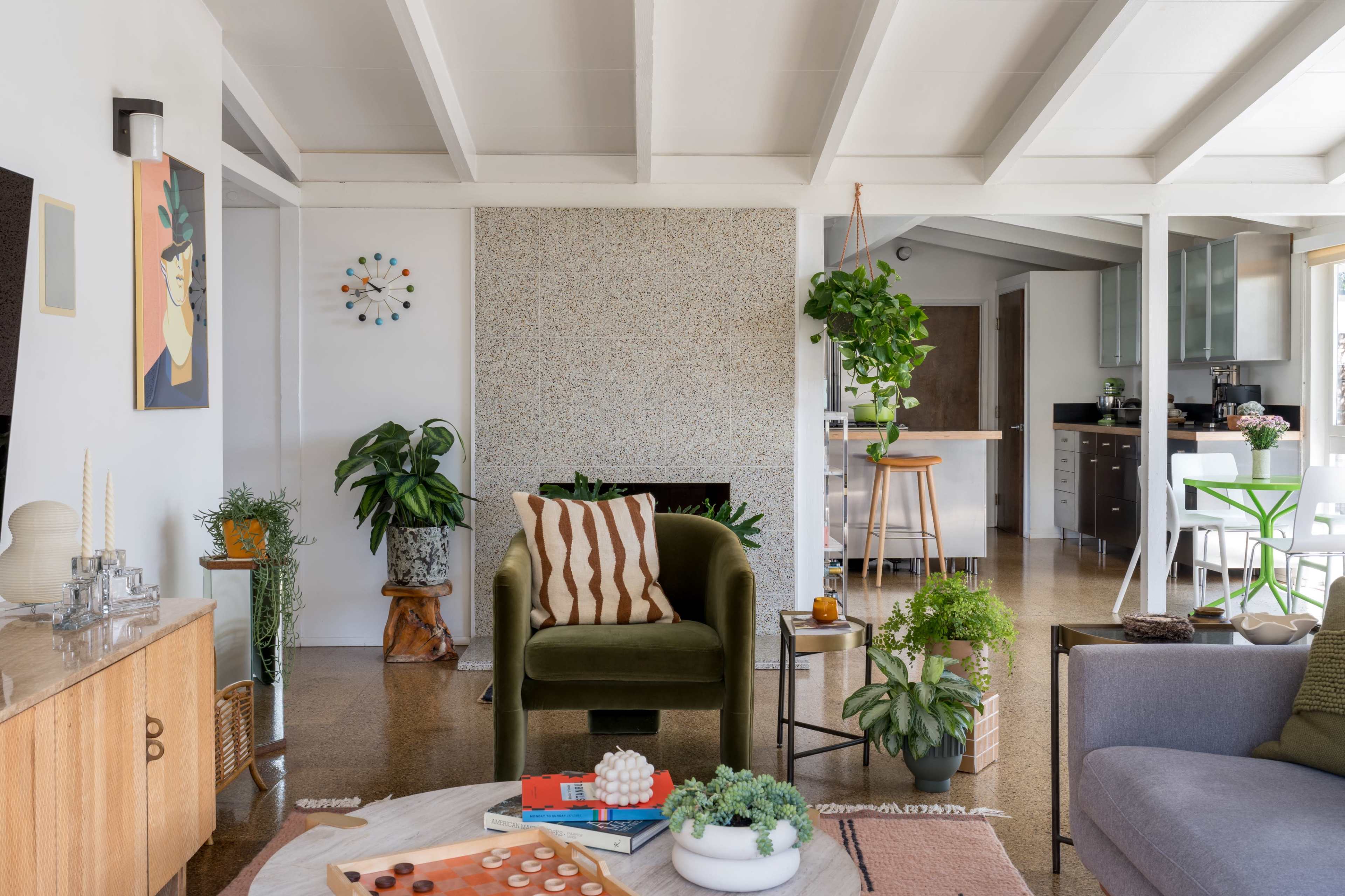 The image shows a stylish living room with a seating area, plants, and a kitchen visible in the background.