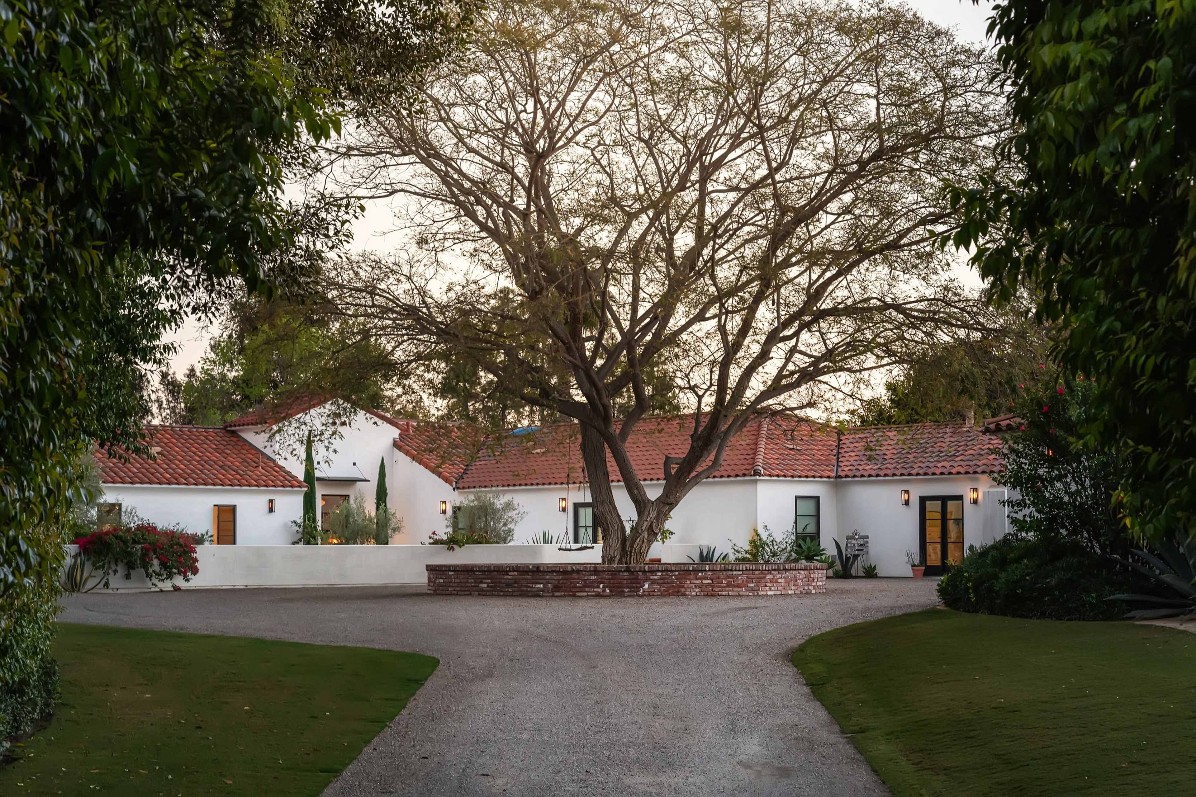 A single-story home with a red-tiled roof and a large tree is situated at the end of a gravel driveway, framed by lush greenery.