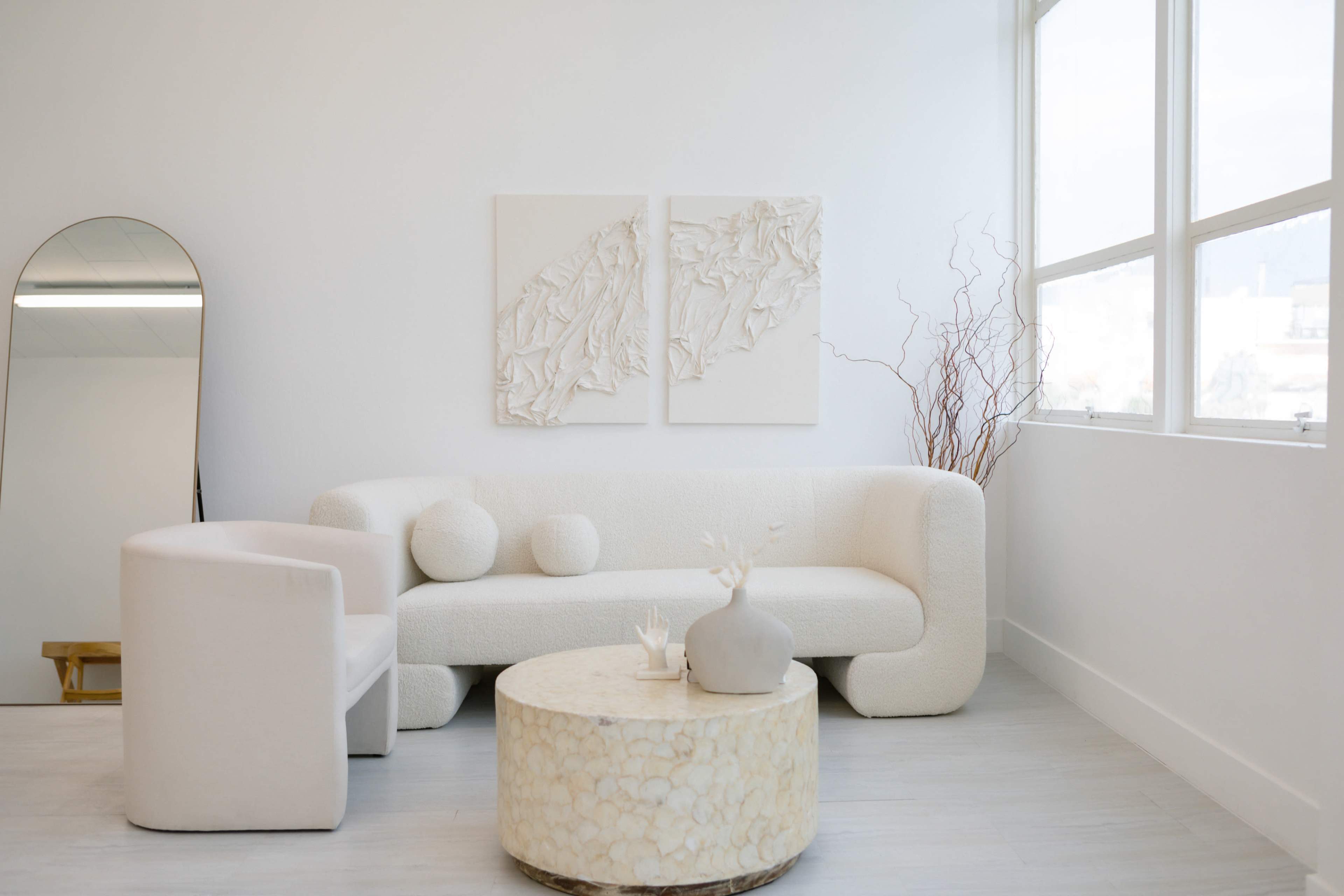 The image shows a minimalist living room with a white curved sofa, a round coffee table, and two textured wall artworks, illuminated by natural light from large windows.