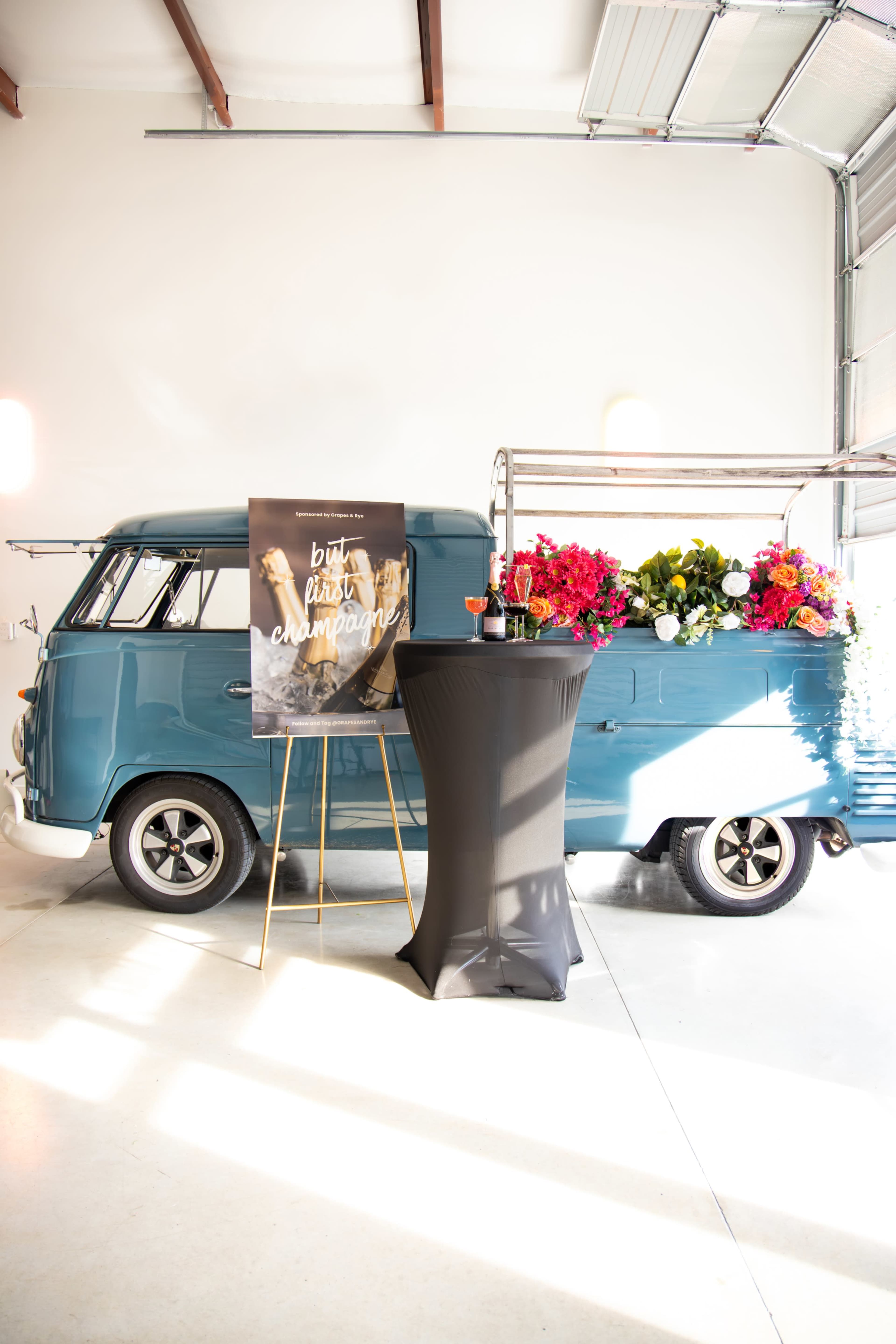 A vintage blue van adorned with vibrant flowers is positioned next to a black cocktail table featuring a sign that reads "but first, champagne" in a well-lit garage space.