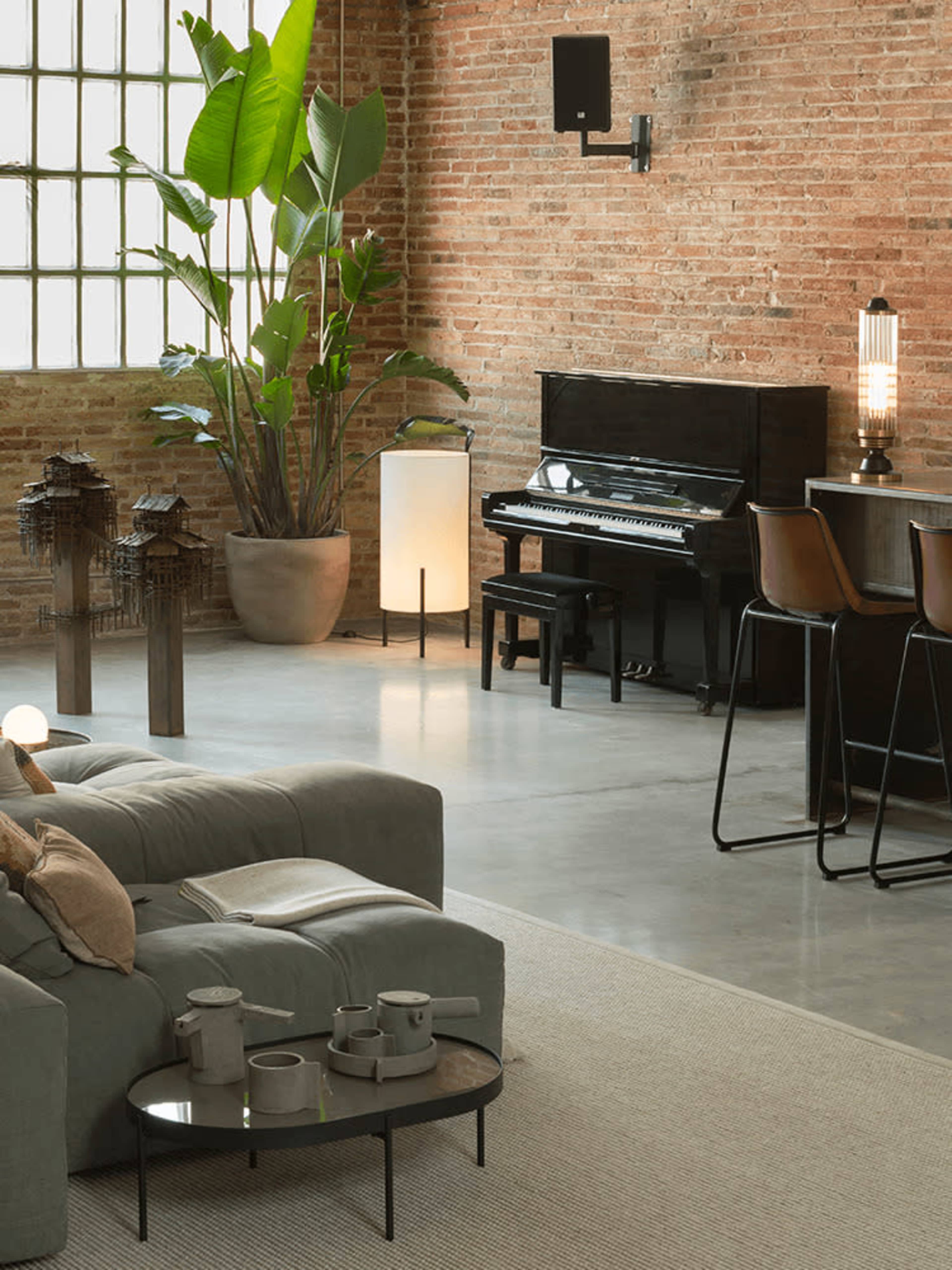 The image features a modern living space with a grey sectional sofa, a black piano, a round coffee table, and a bar area with three stools, all set against a backdrop of exposed brick walls and a large window.