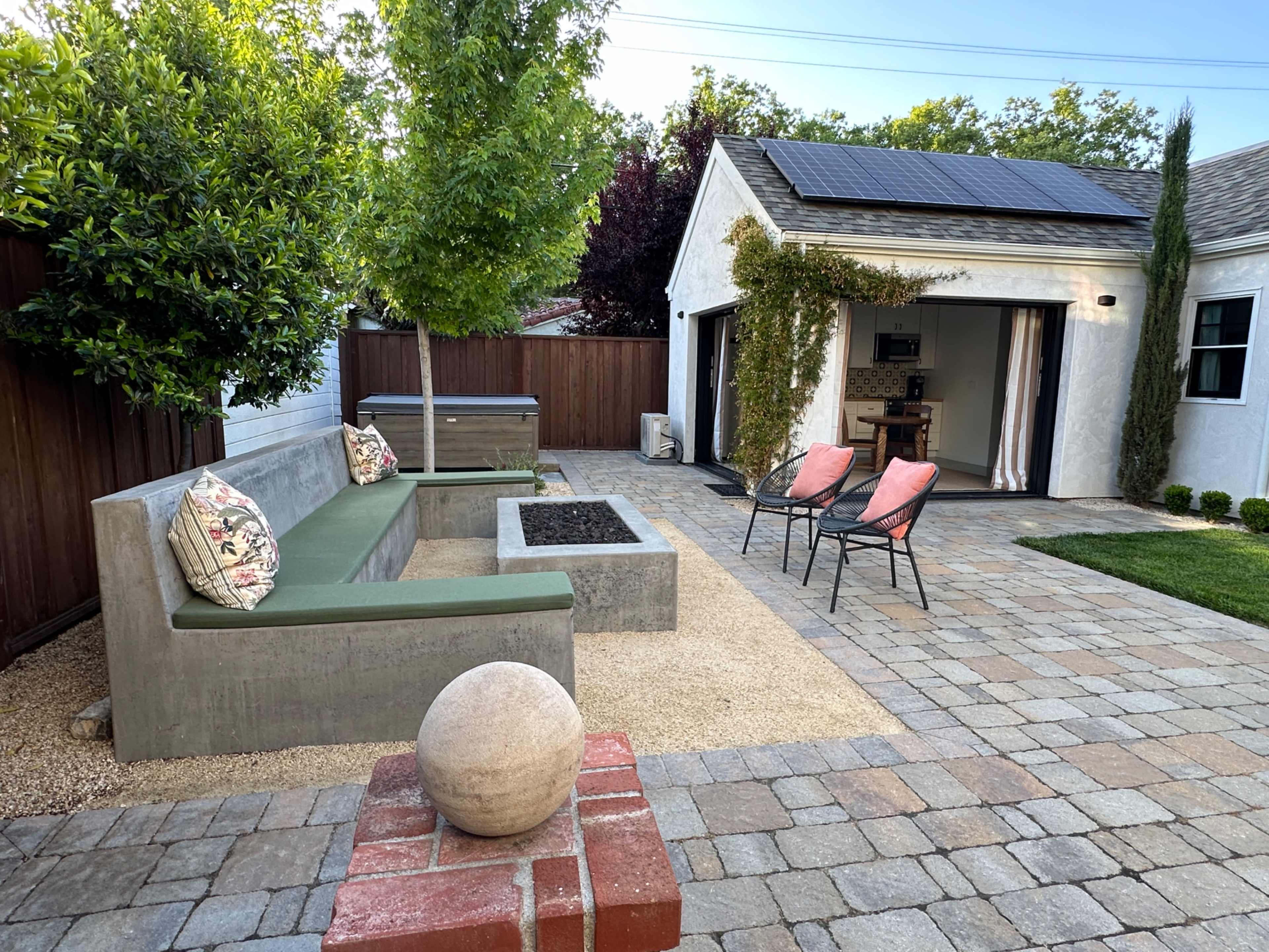The image shows a modern backyard featuring a concrete seating area, a fire pit with a circular stone, and two red chairs, surrounded by landscaped greenery and a small structure.