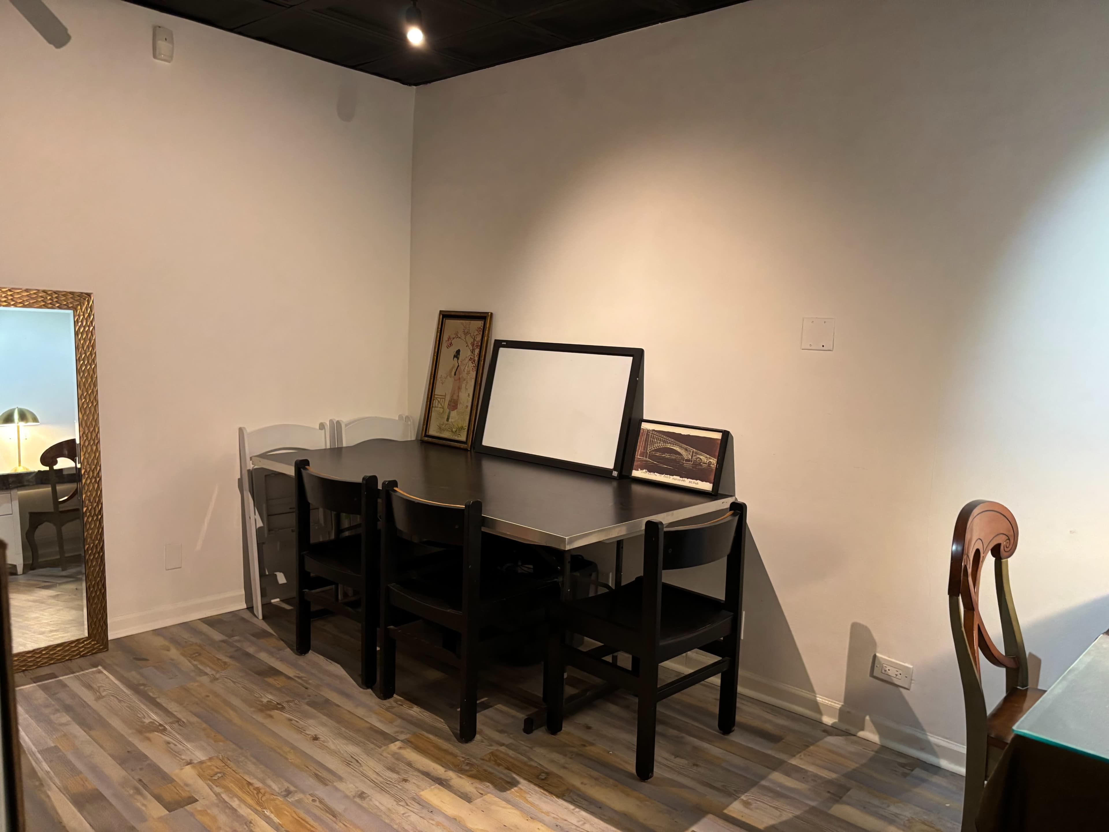 A dark wooden table with black chairs and two framed pictures is placed against a white wall in a room with wooden flooring.