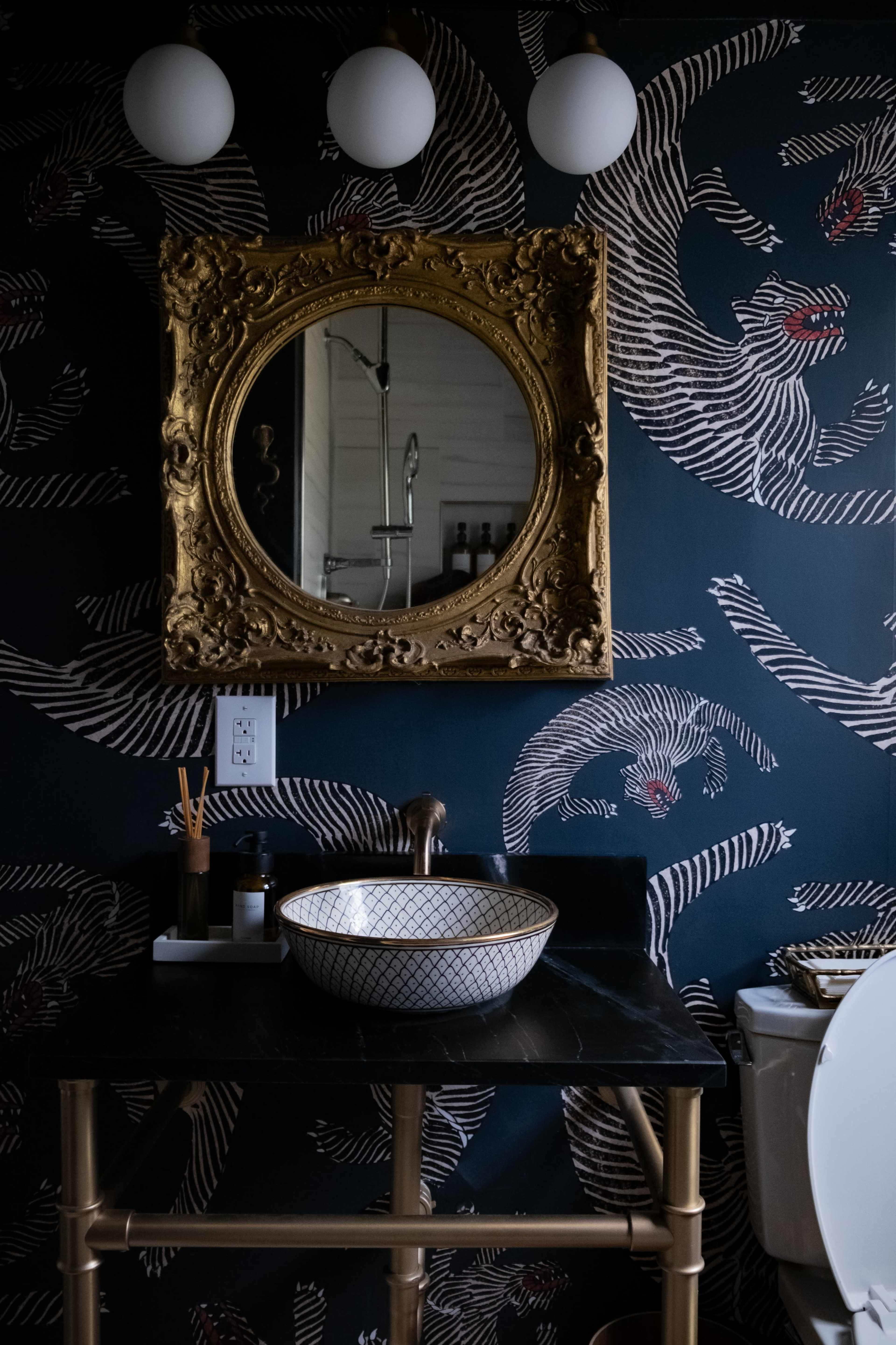 A bathroom featuring a decorative gold-framed mirror above a dark marble sink with a patterned bowl, alongside a toilet and patterned wallpaper.