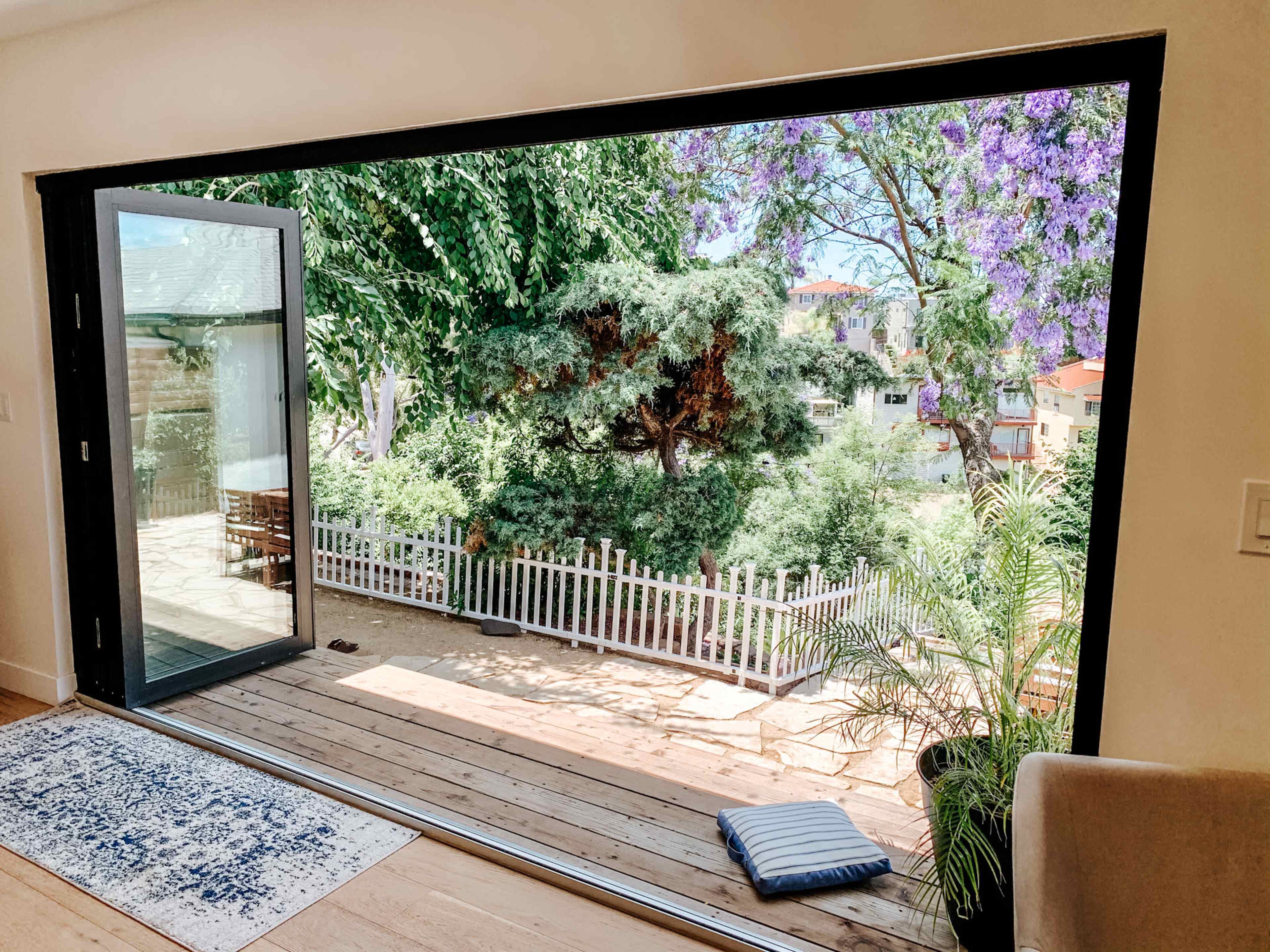A large sliding glass door opens to a wooden deck surrounded by greenery and a white picket fence.