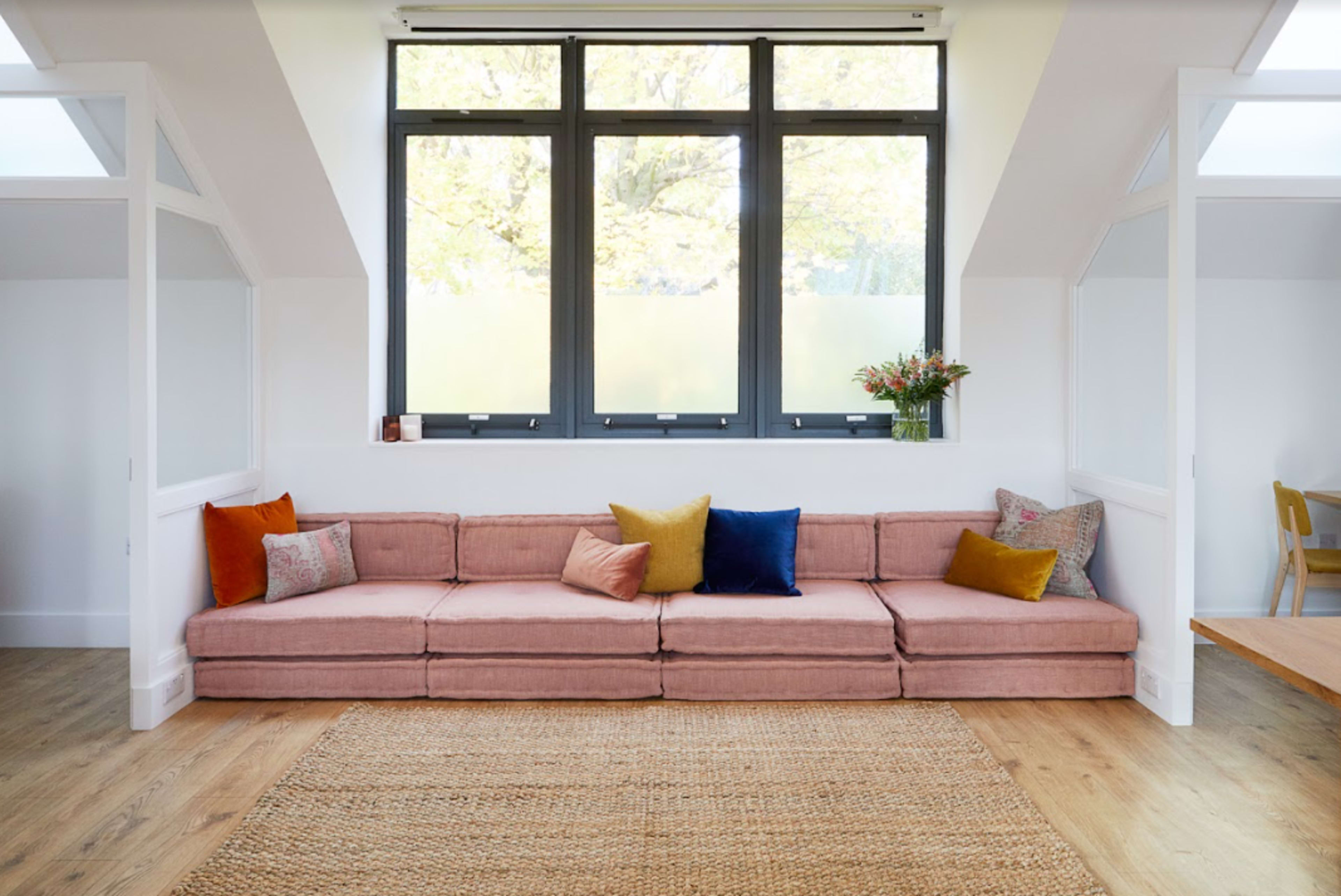 A modern living room featuring a pink sectional sofa with colorful pillows, a woven rug, and large windows allowing natural light to fill the space.