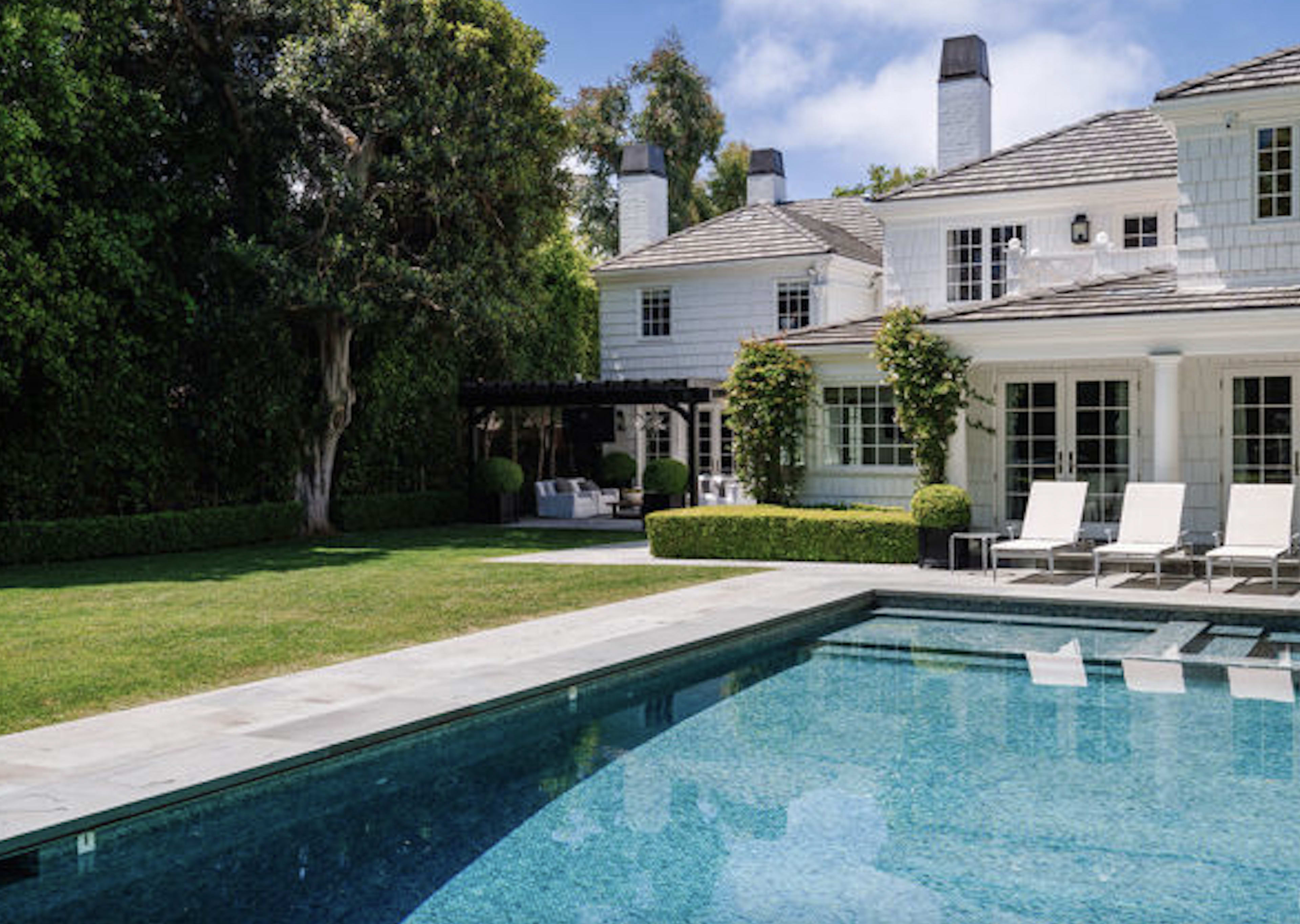 The image shows a modern house with a large swimming pool in the foreground, surrounded by landscaped gardens and patio furniture.