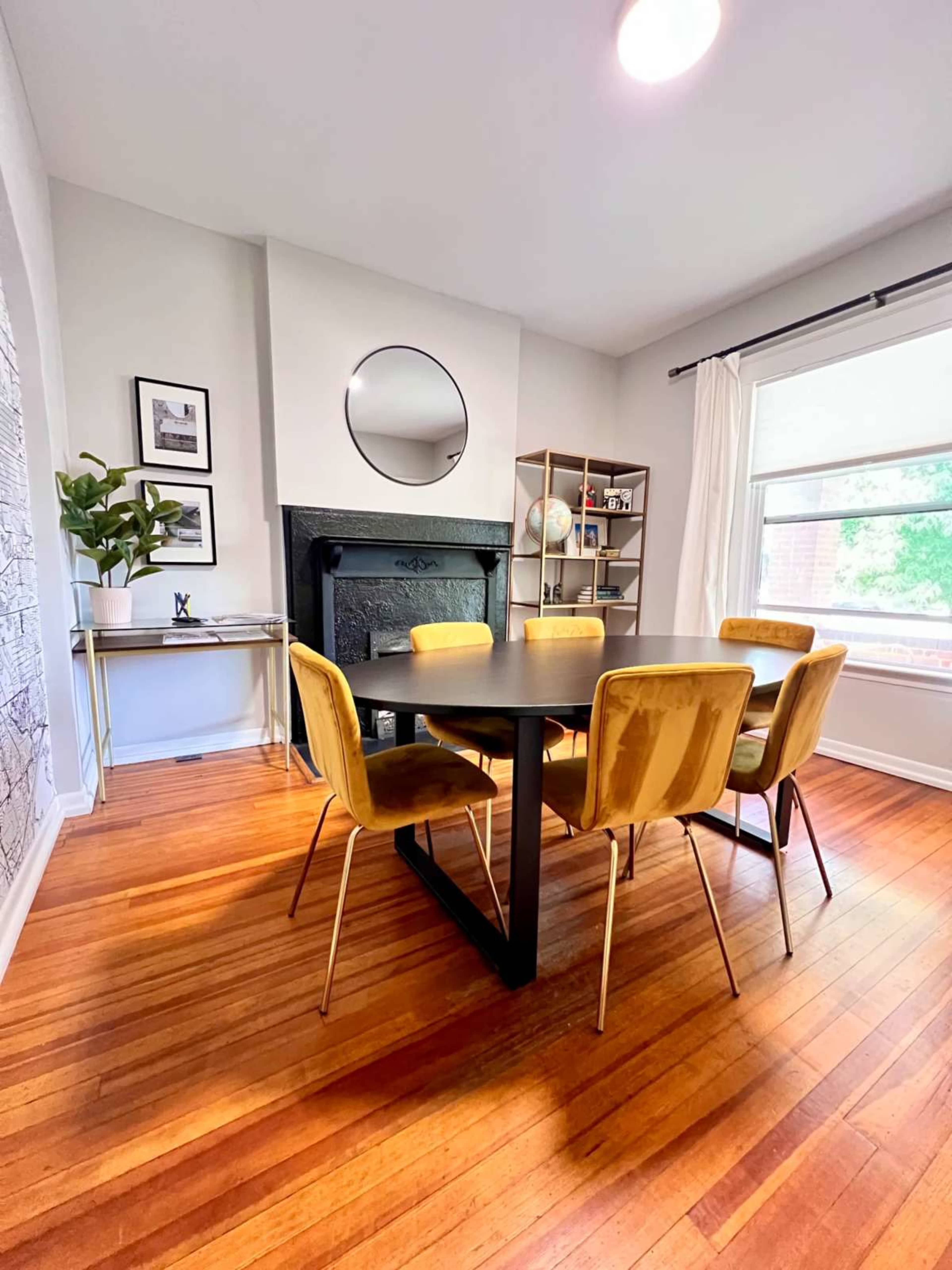 A dining area features a round black table surrounded by six yellow chairs, with a mirror and a bookshelf on the walls.