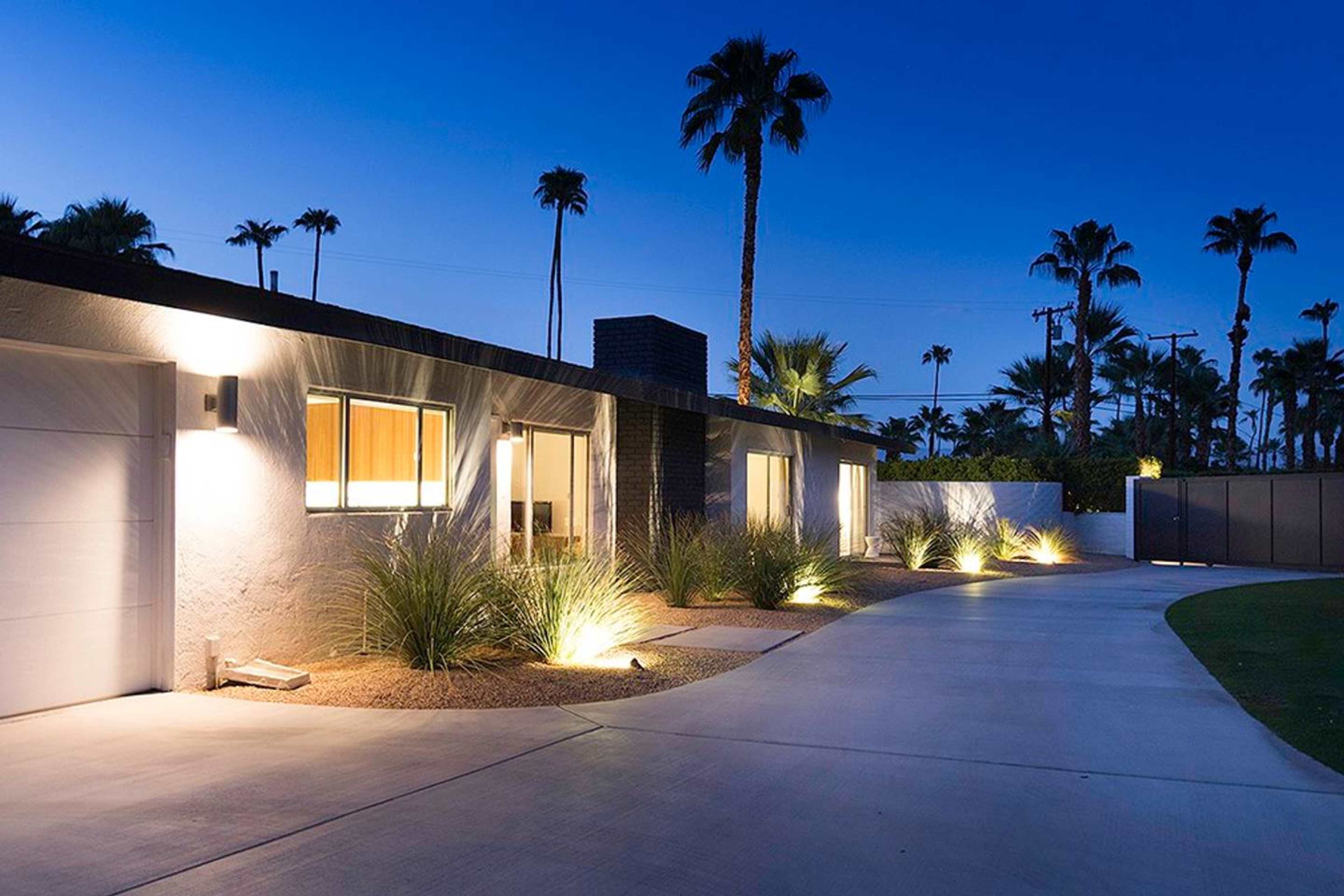 A modern single-story house with a landscaped driveway is illuminated by outdoor lighting, surrounded by palm trees at dusk.