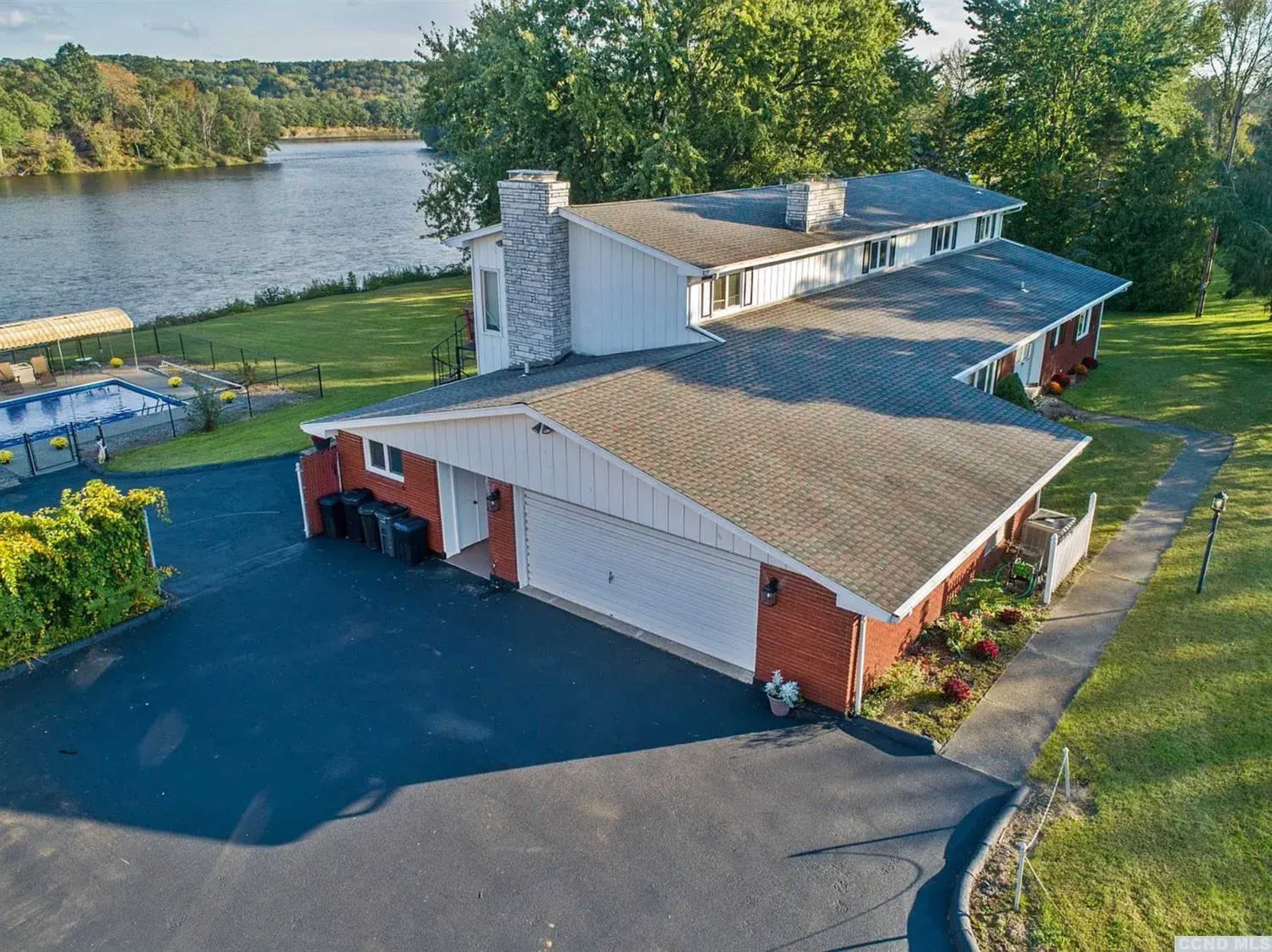The image shows a large, two-story house situated by a river, with a swimming pool area and well-maintained landscaping.