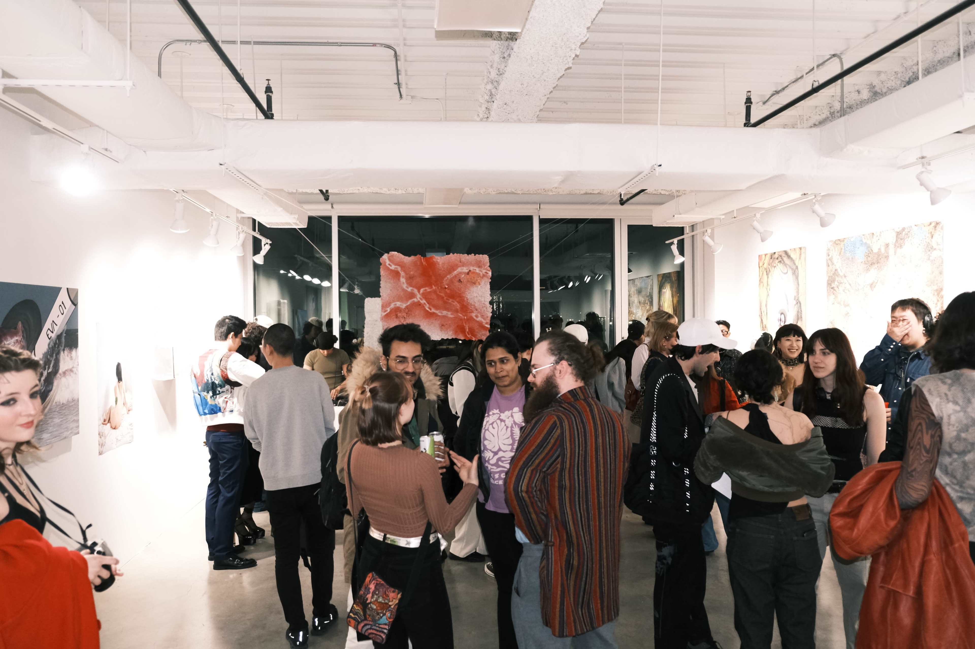 A crowd of people gathers inside a gallery, admiring artwork displayed on the walls while socializing and holding drinks.