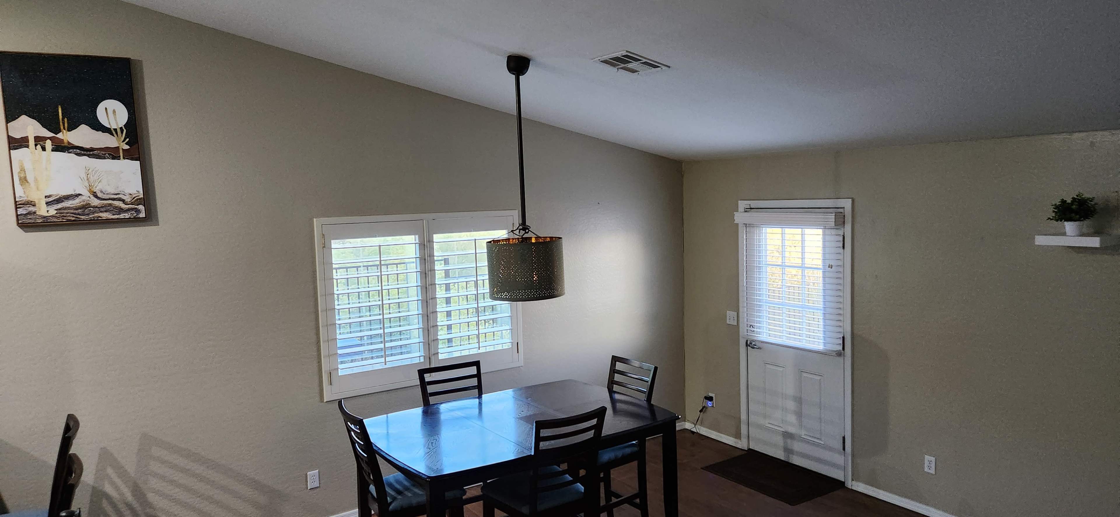 A dining area features a table with four chairs, a pendant light, and a door leading outside, with a wall art piece hanging above the table.