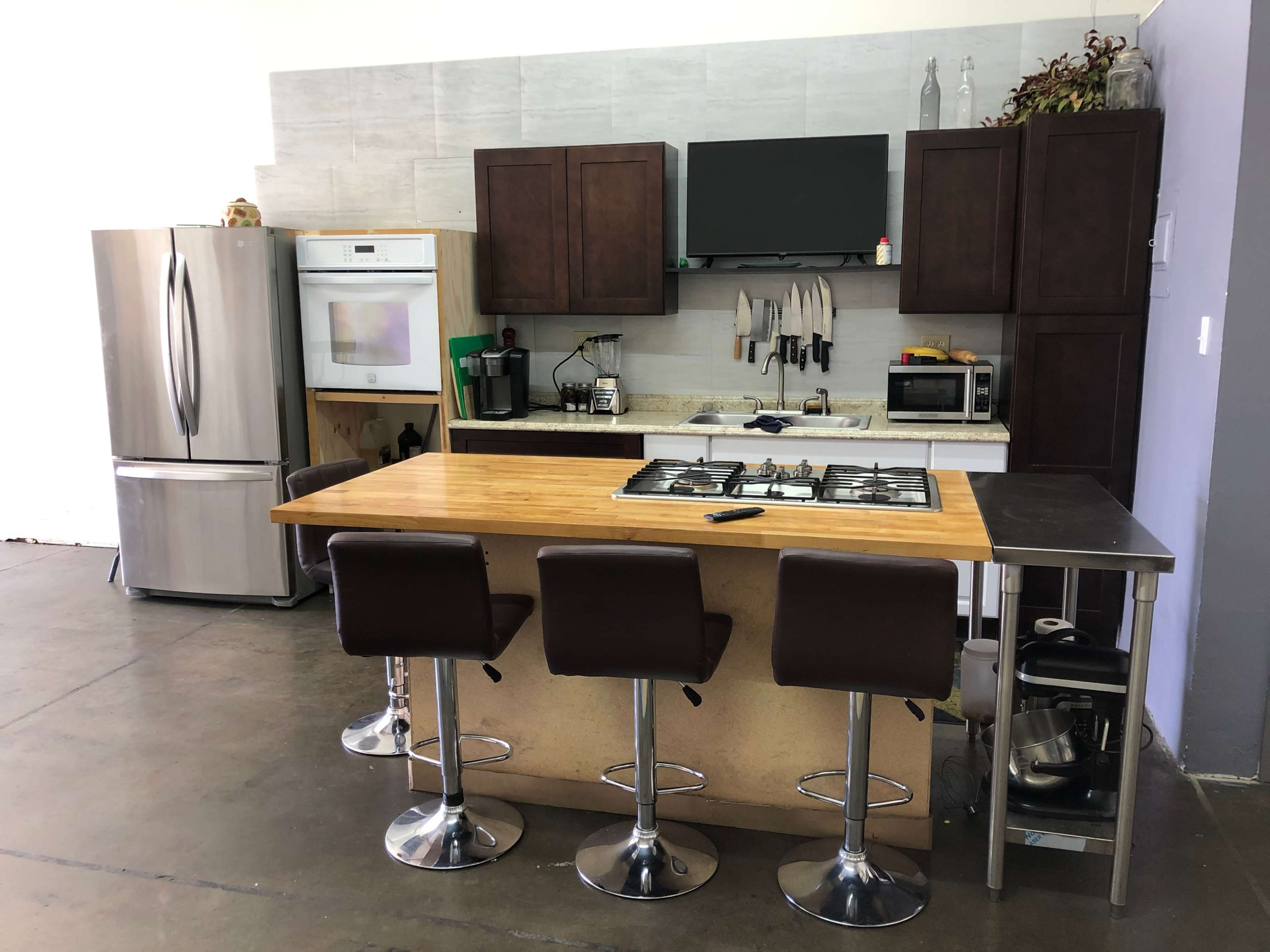 The image shows a modern kitchen featuring dark wooden cabinets, a stainless steel refrigerator, a gas stove on an island with three barstools, and various kitchen appliances.