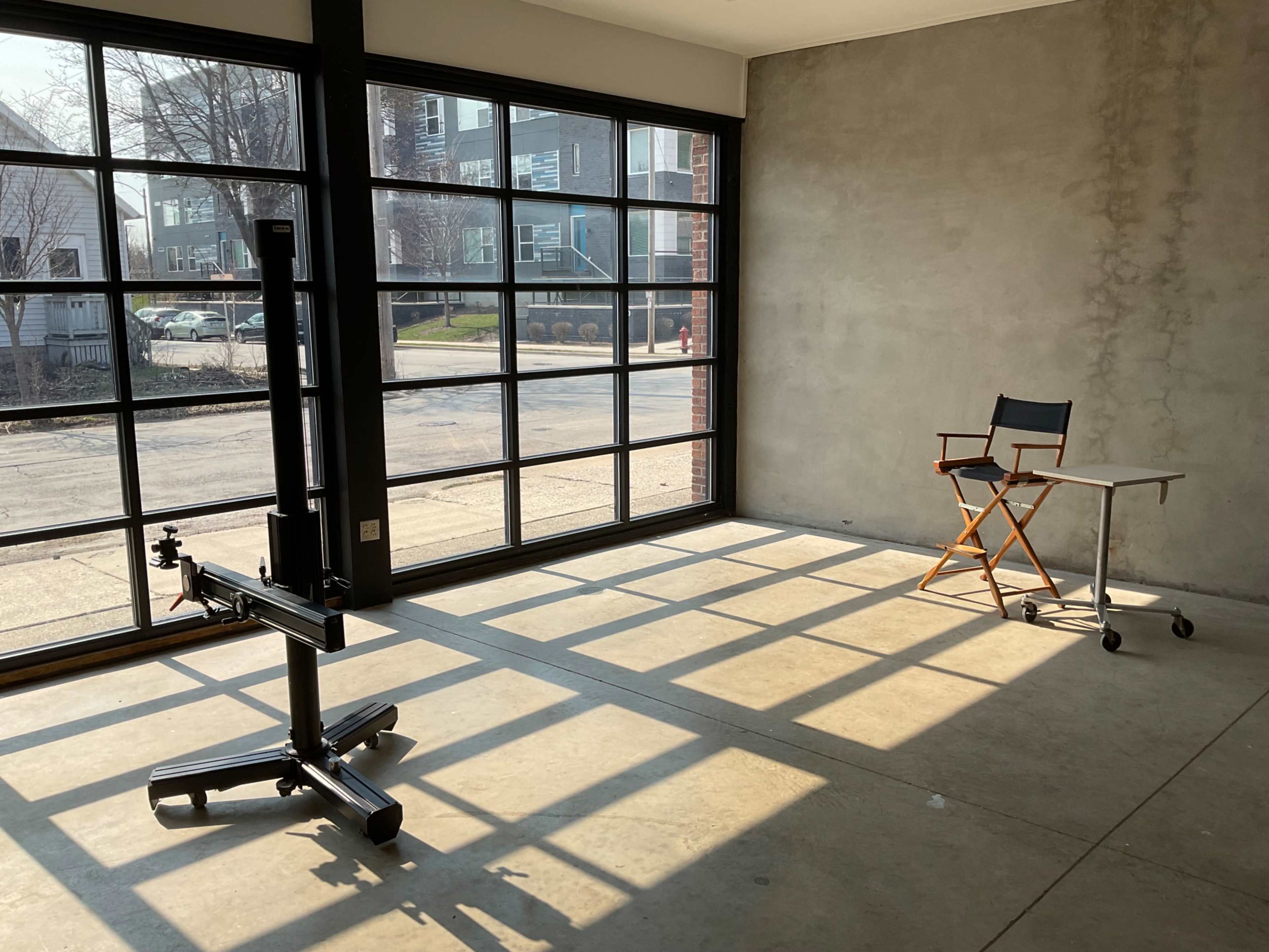 A sunlit room with large windows, featuring a black cart on wheels and a folding chair next to a small table.