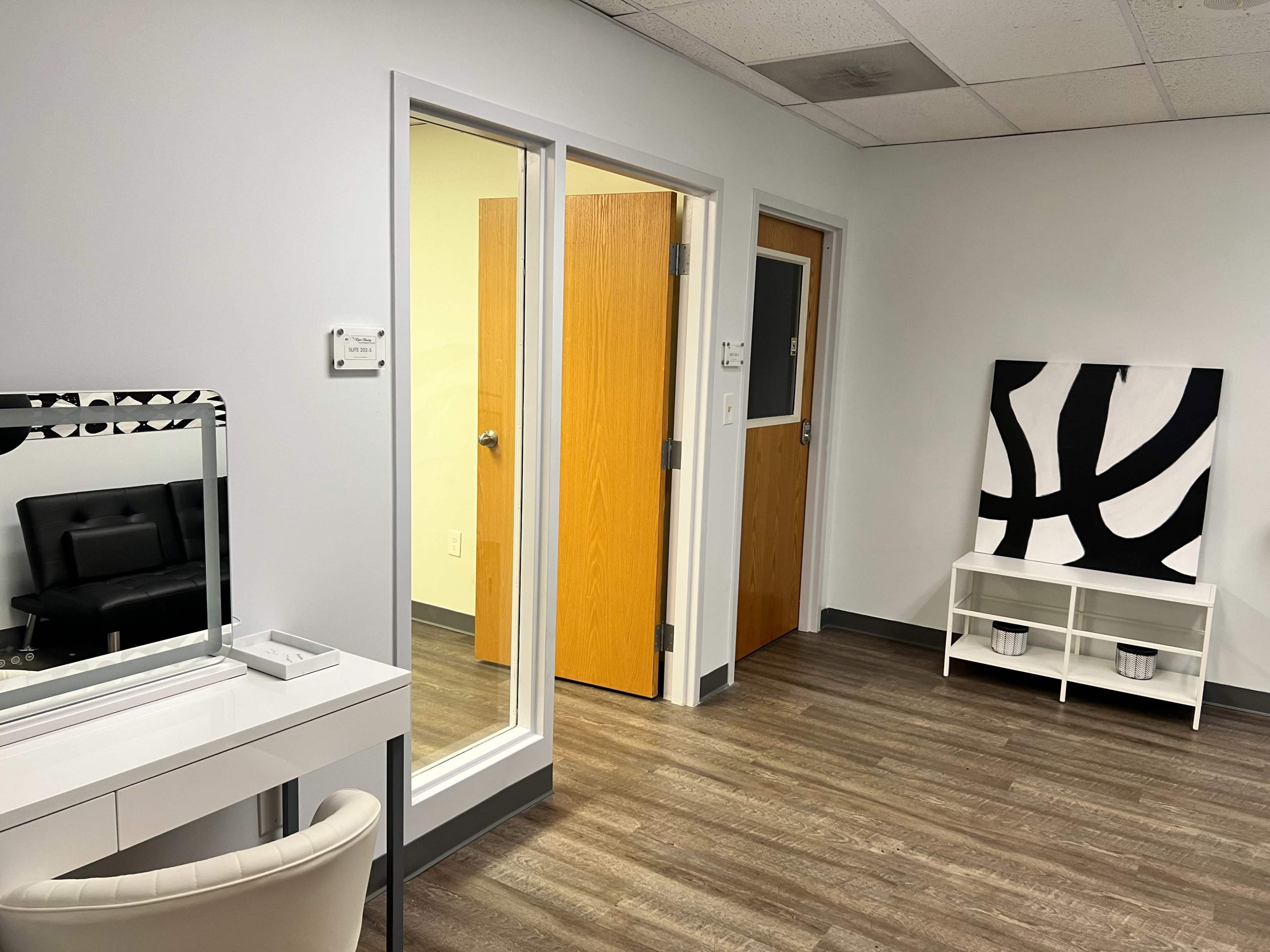 The image shows a brightly lit interior of an office with a mirror and a chair on one side, doors leading to other rooms, and a large abstract art piece on a shelf against the wall.