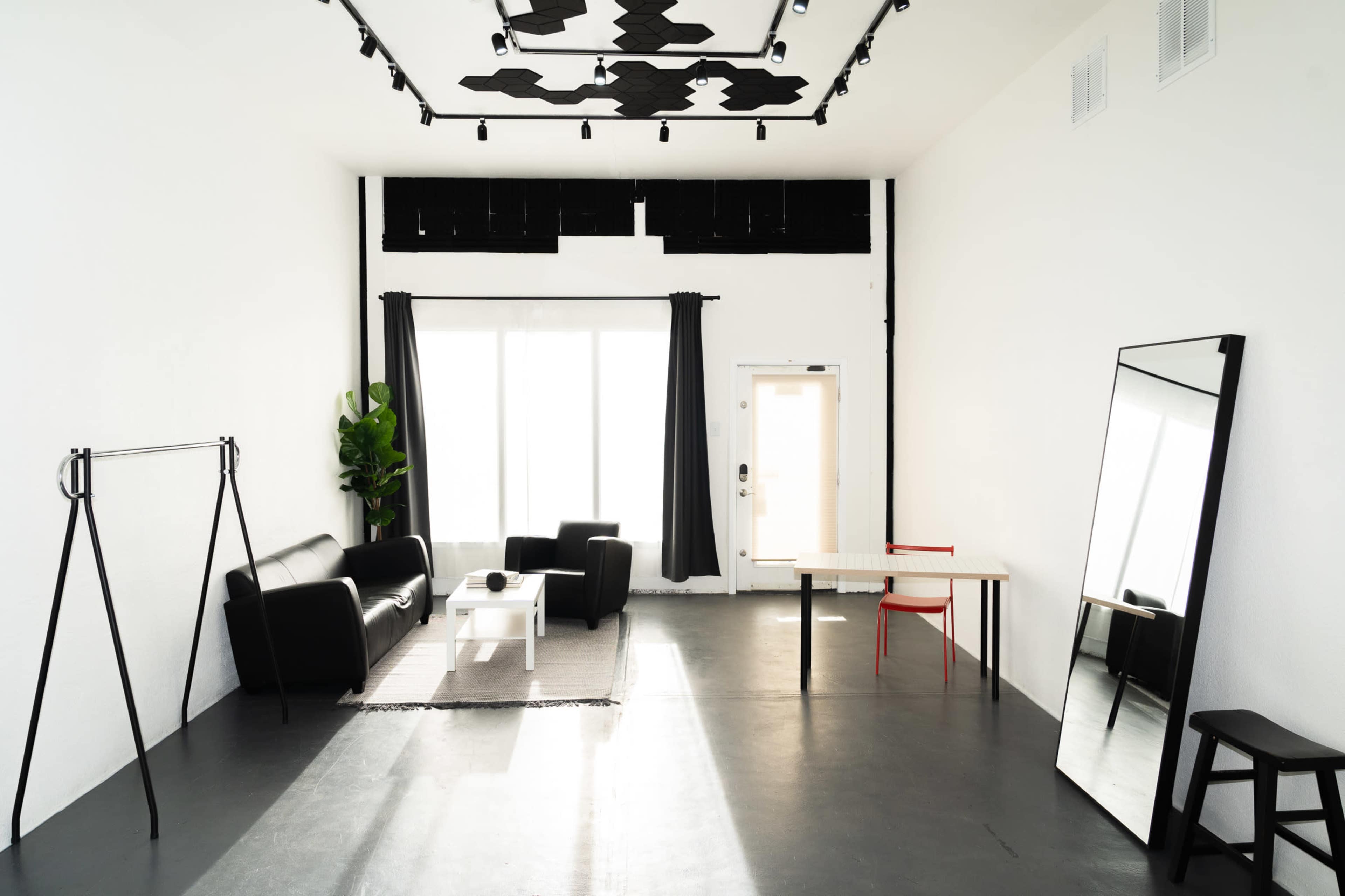 A minimalist room with a black sofa, a white coffee table, a large mirror, and a table with a red chair, illuminated by natural light from a large window.