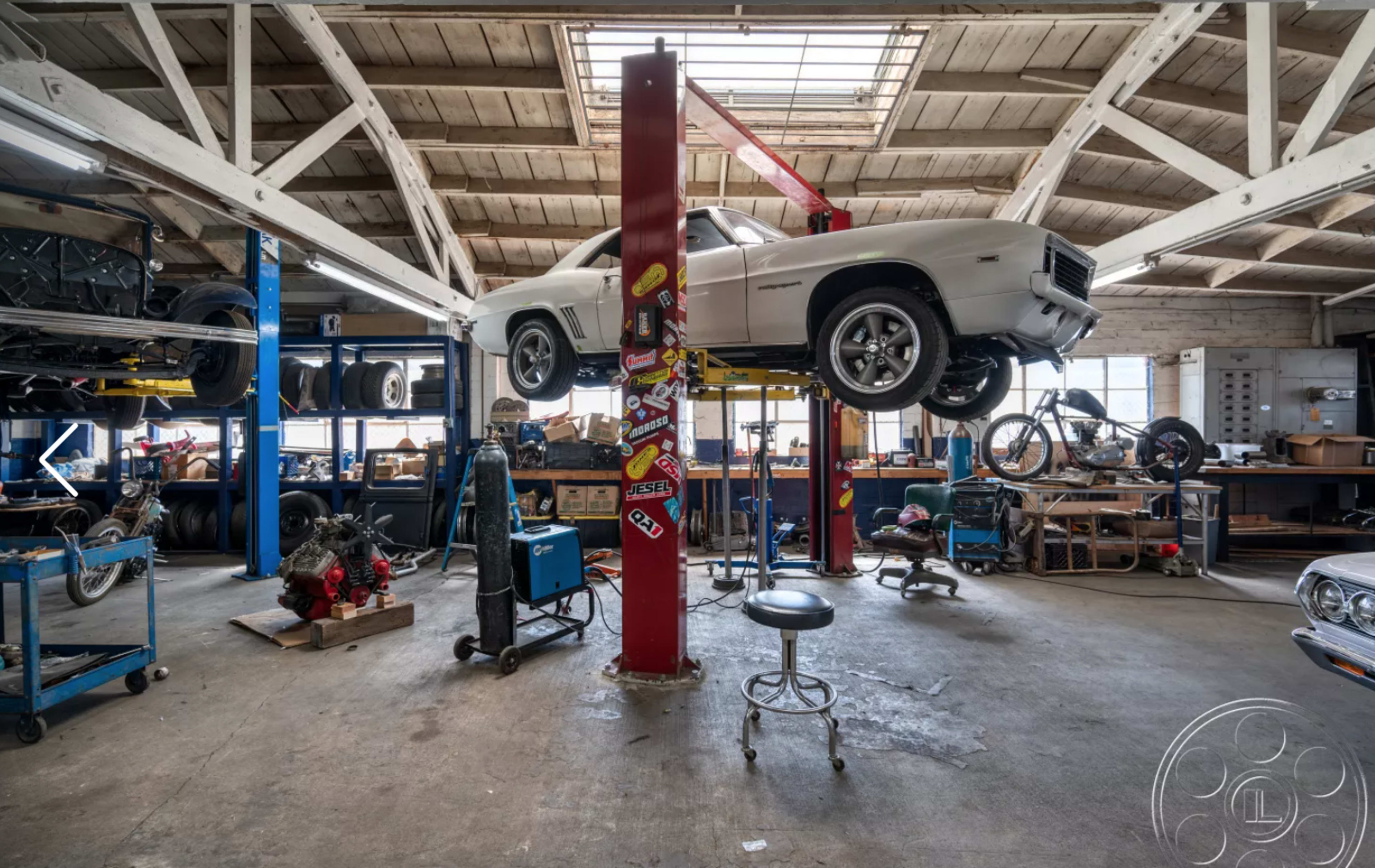An automotive workshop features a white car elevated on a lift, surrounded by tools and parts.