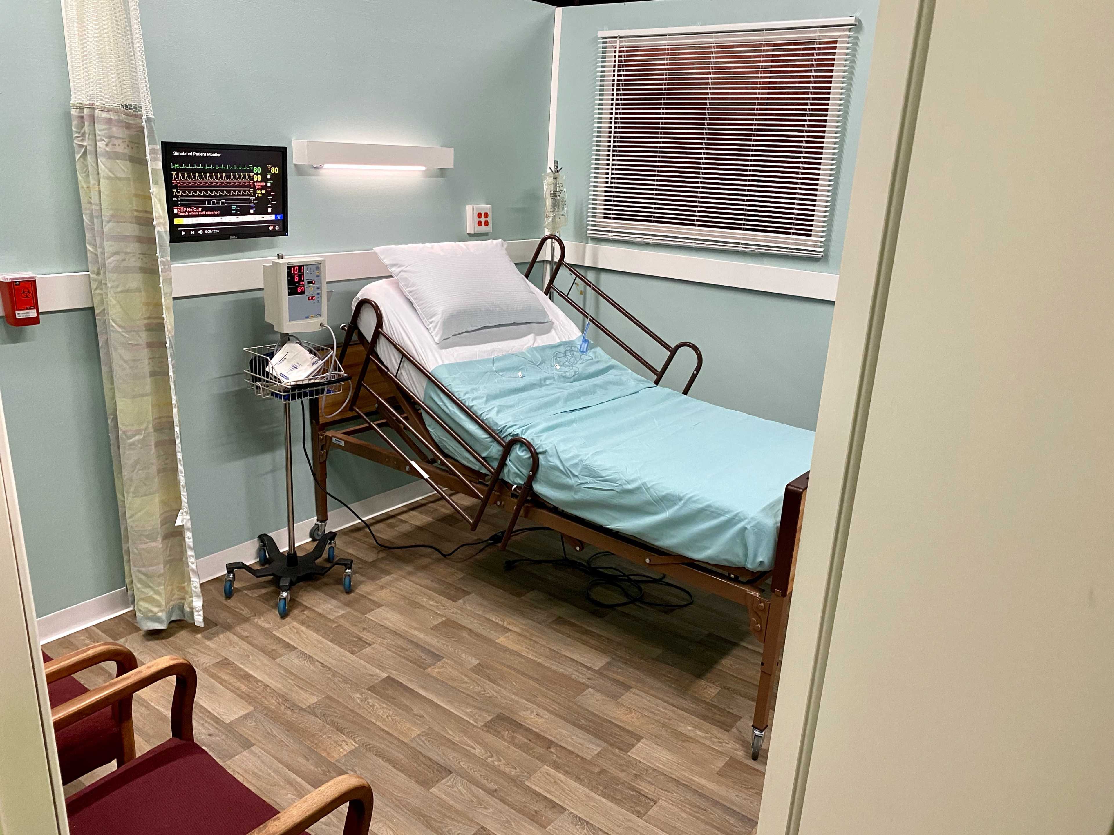 A hospital room featuring a medical bed with a light blue sheet, a wall-mounted monitor, and two chairs positioned near the entrance.