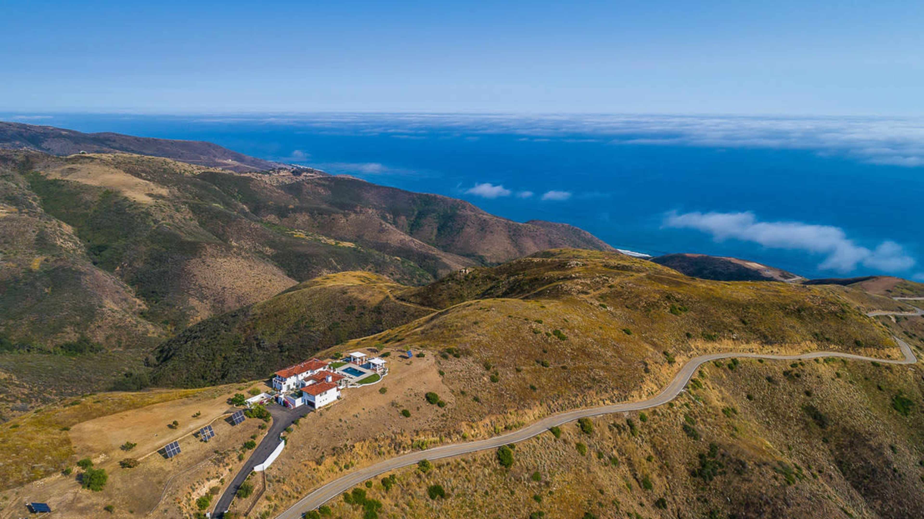 A large villa is situated on a hillside overlooking a blue ocean and rugged coastline.