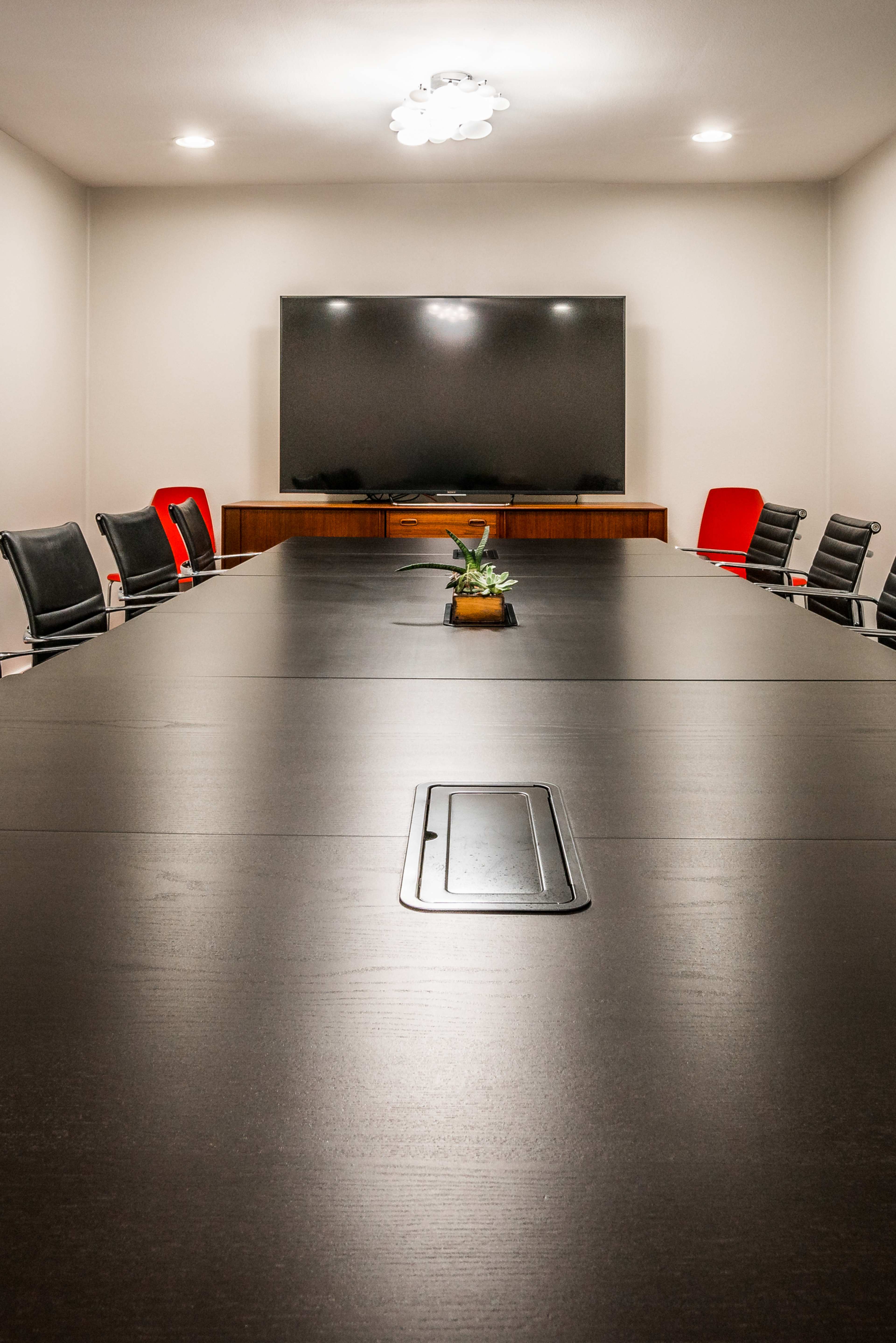 A large conference room features a long black table with several black and red chairs, a flat-screen TV on the wall, and a small plant centerpiece.