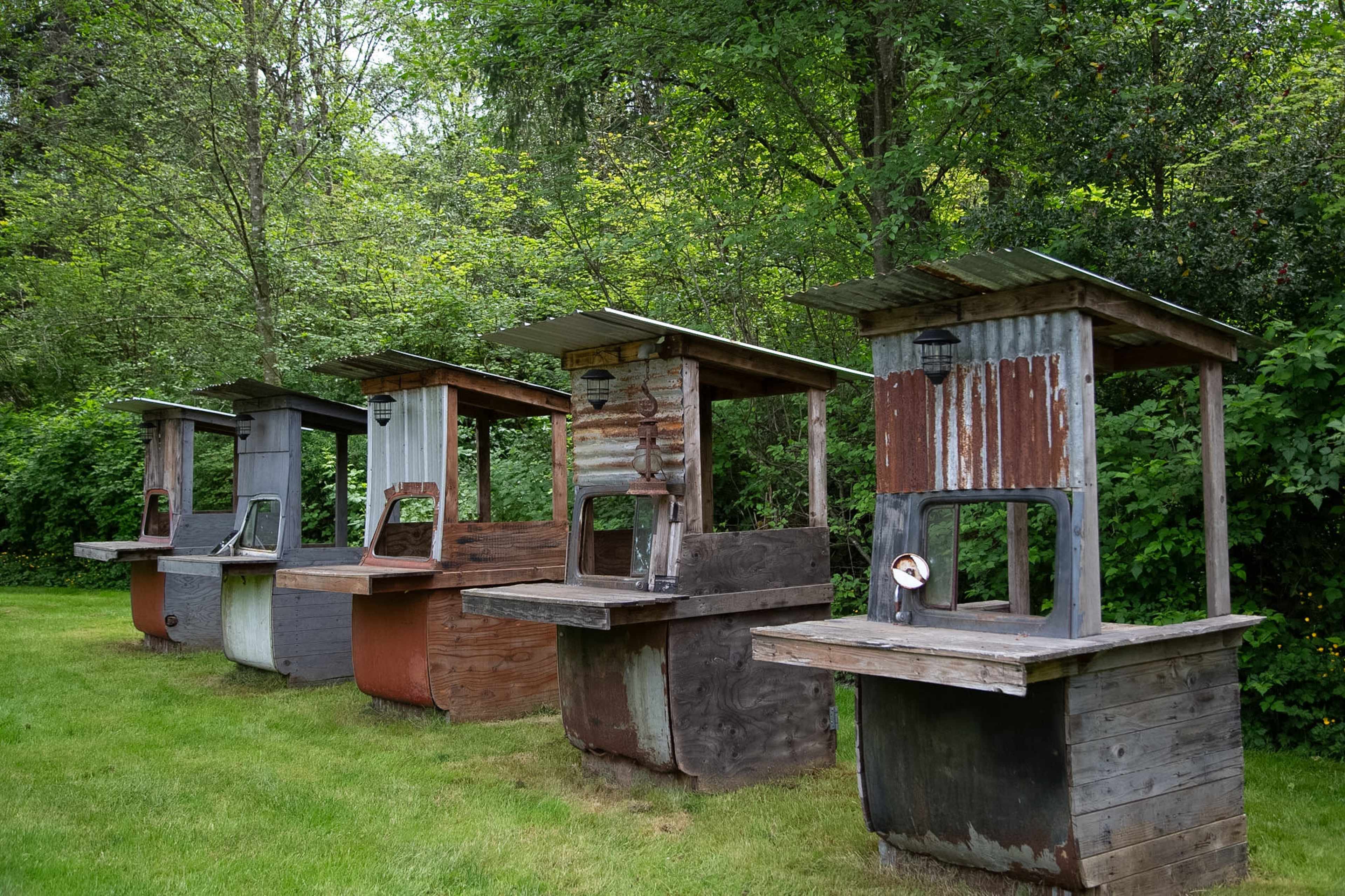 The image shows five rustic, open-air stalls with corrugated metal roofs and wooden bases arranged in a row, surrounded by greenery.