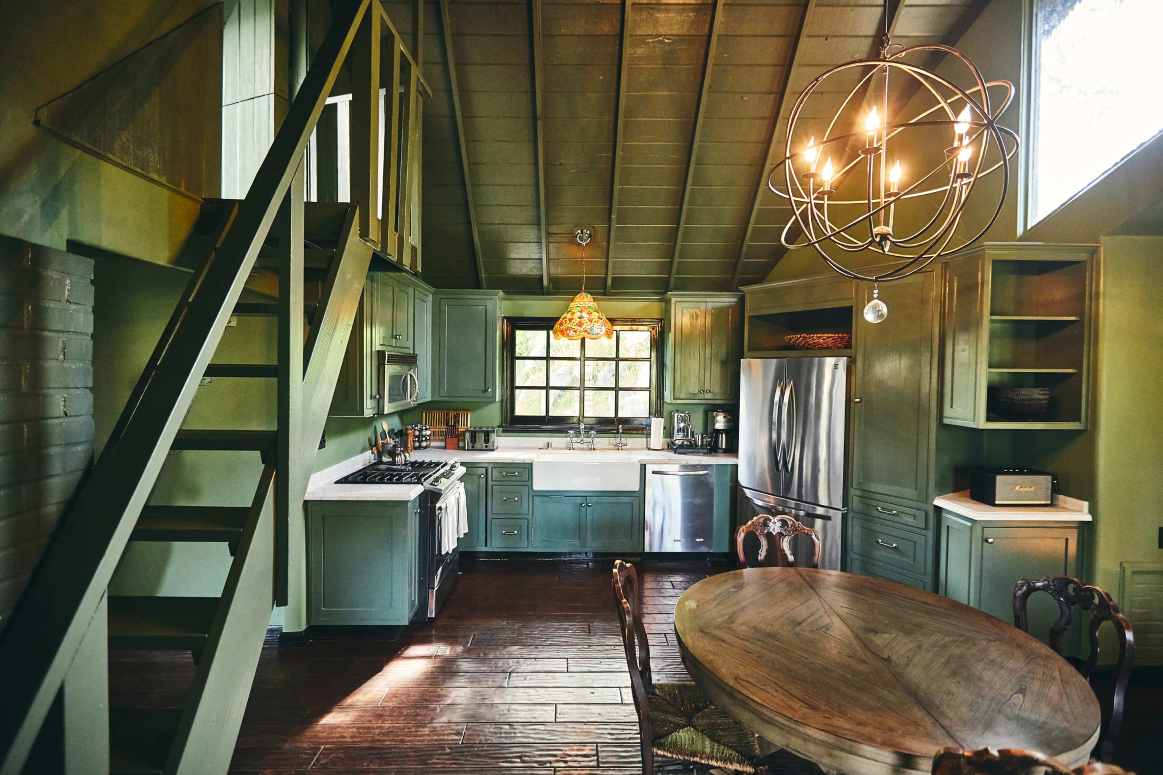 The image shows a rustic kitchen and dining area with green cabinetry, hardwood flooring, and a chandelier.