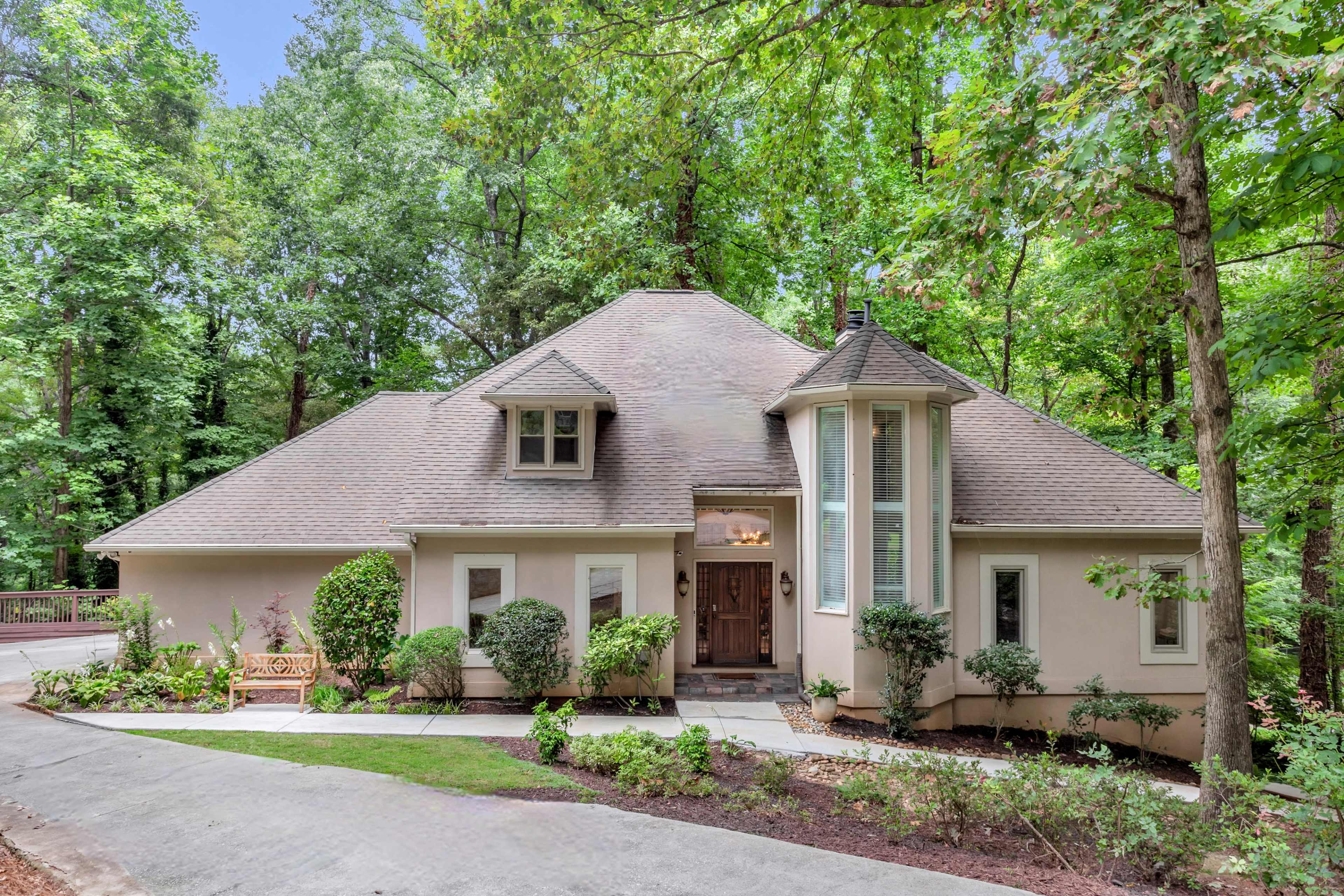 A single-story house with a unique roof design is set among tall trees and landscaped greenery.