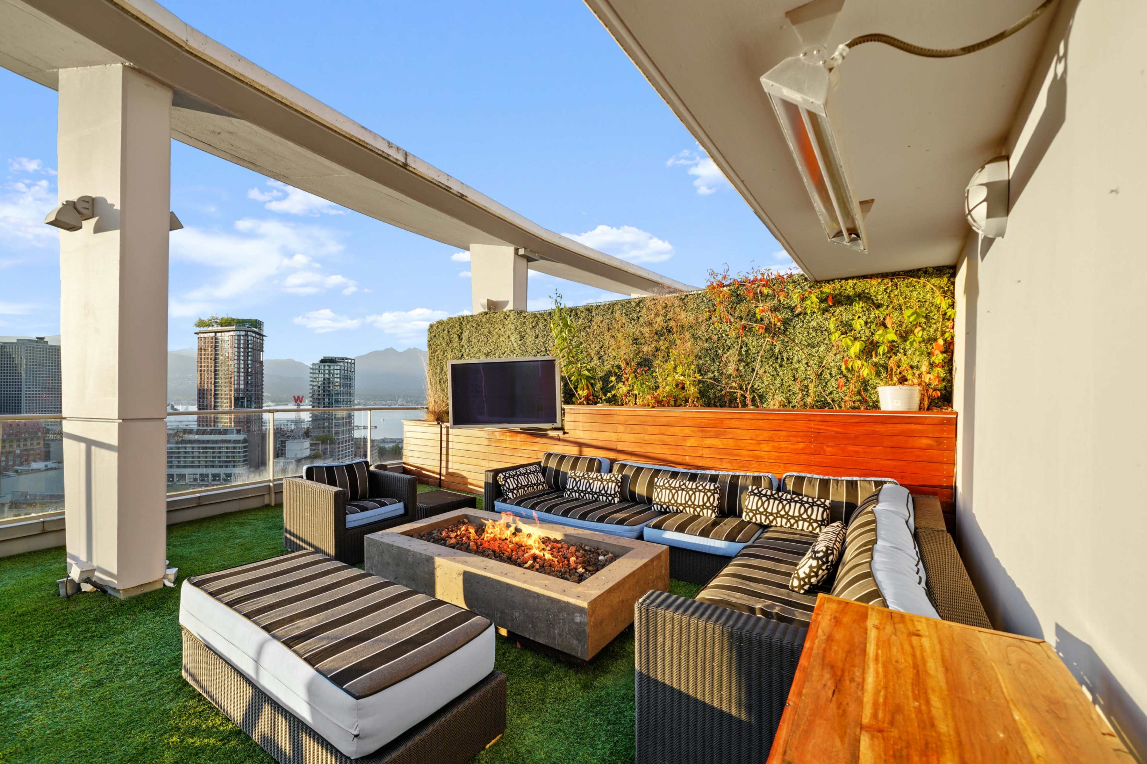 The image shows a rooftop terrace featuring a modern seating area around a fire pit, with a backdrop of city buildings and mountains.