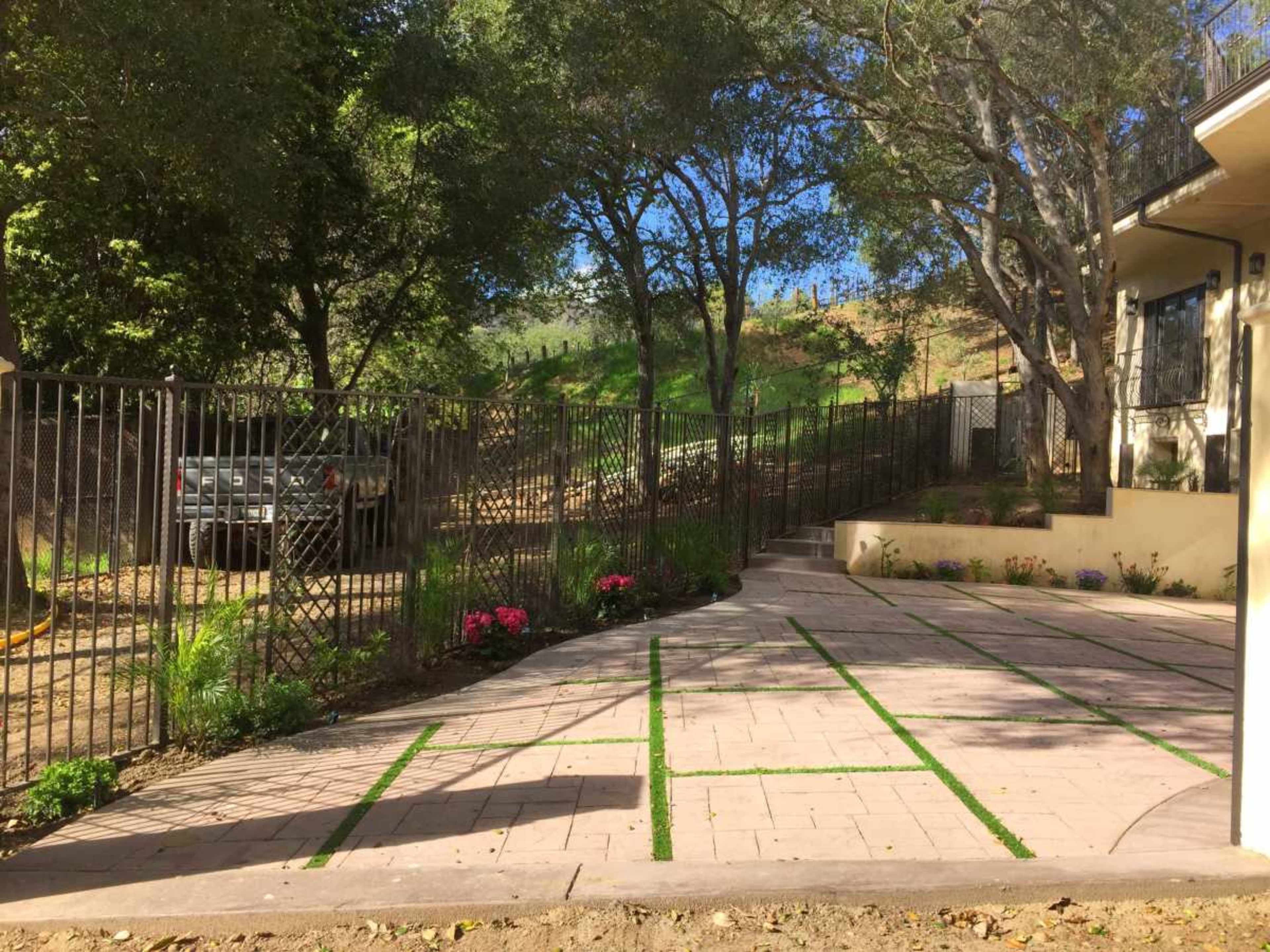 The image shows a paved outdoor area with flower beds and a fence, surrounded by trees and greenery.