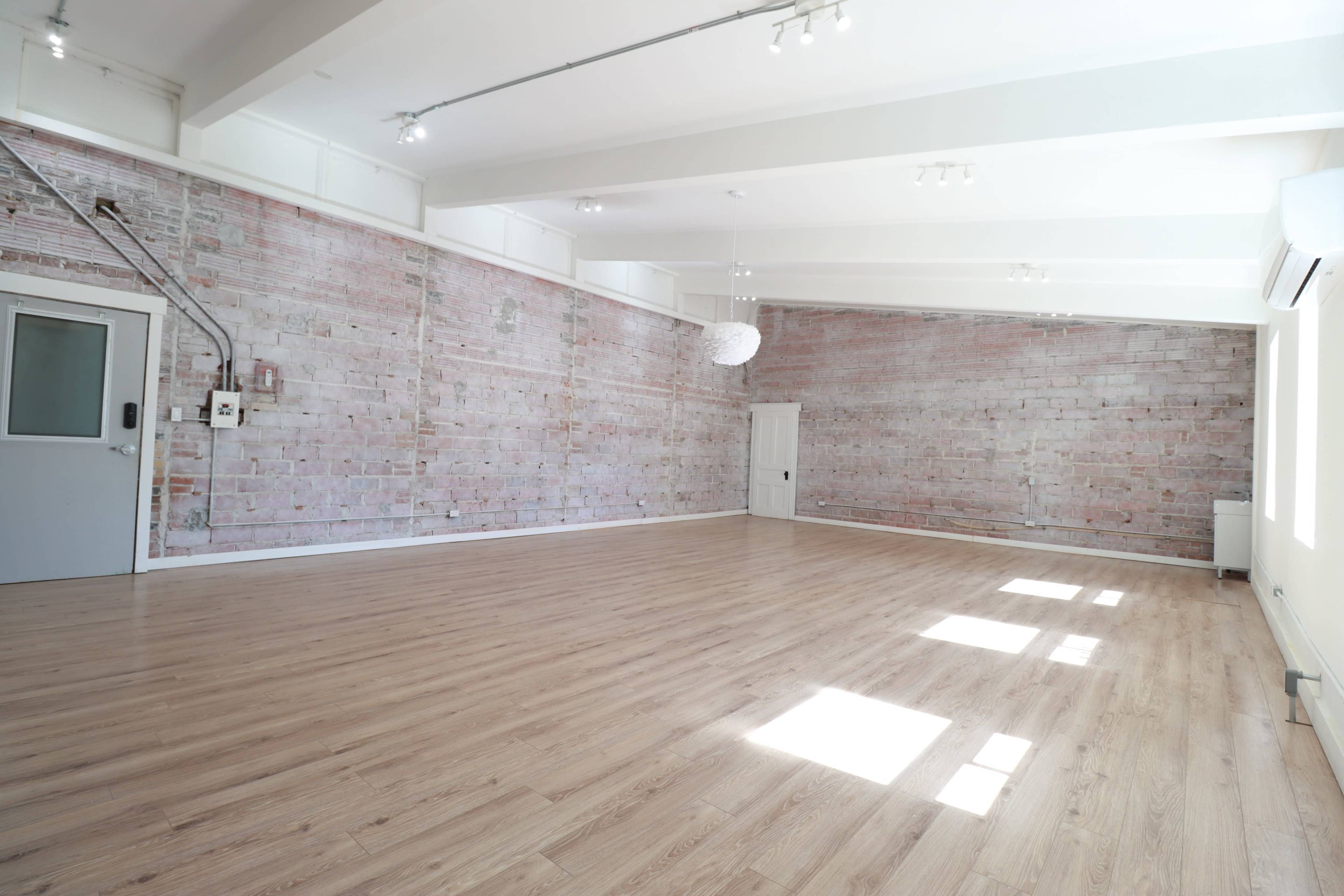 A spacious room with light wood flooring and exposed brick walls, illuminated by natural light from several windows.