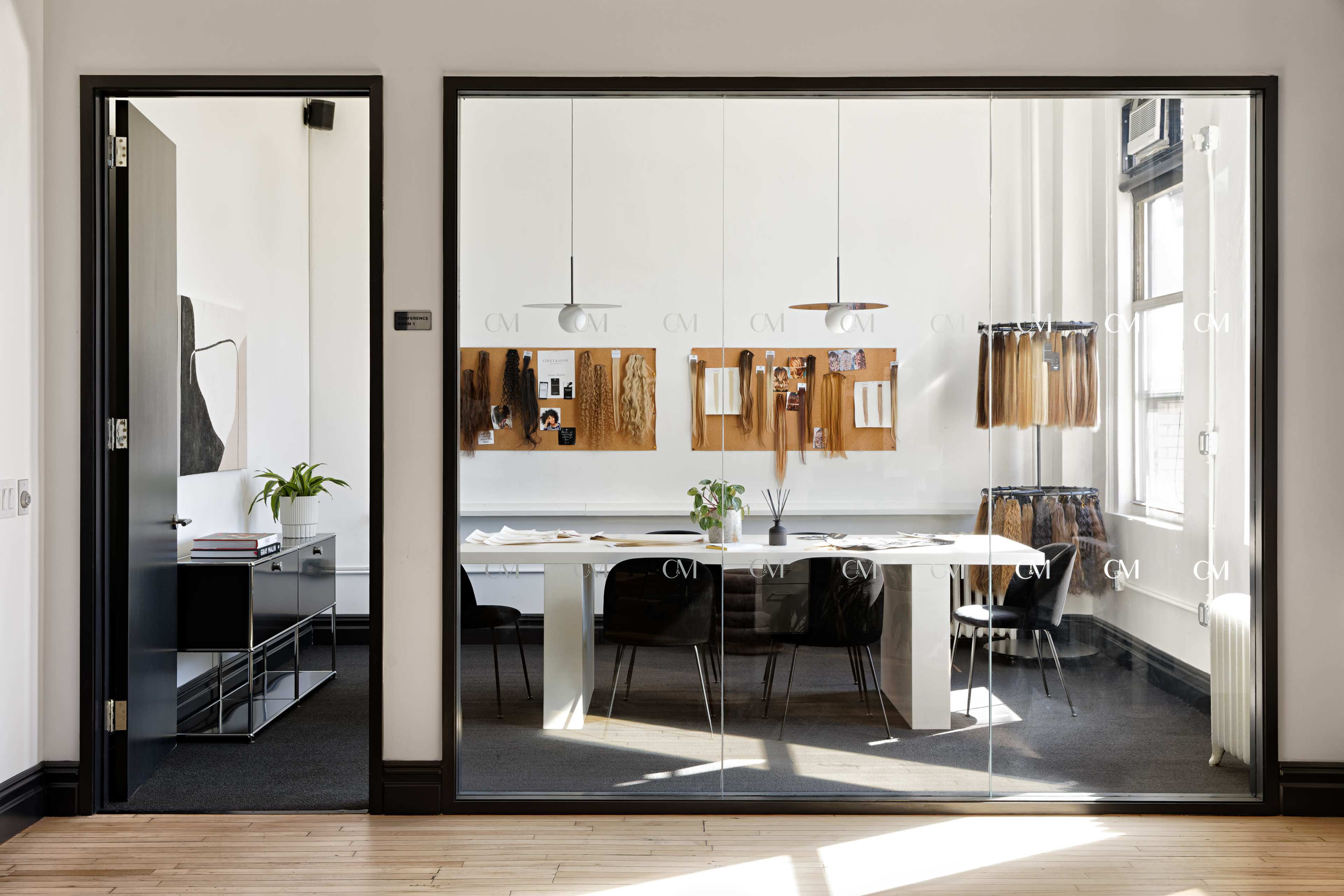 A glass-walled office features a long white table surrounded by black chairs, with wall-mounted boards displaying various materials and a plant on the table.