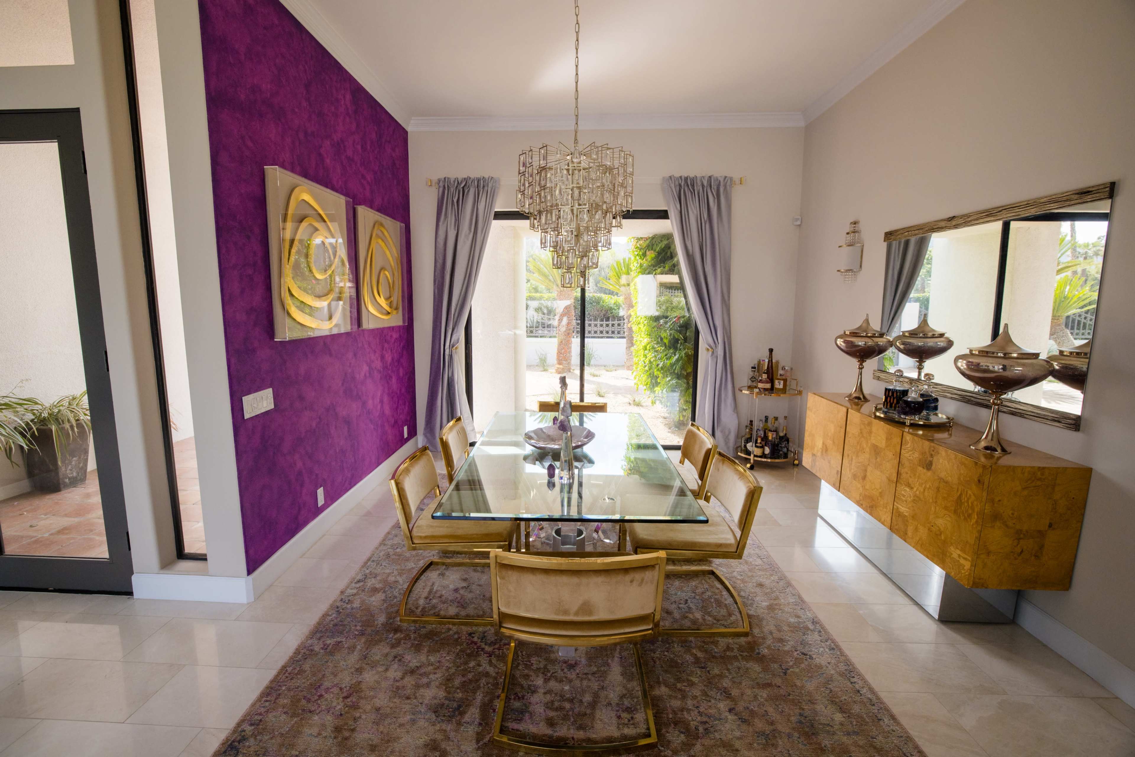 A modern dining room with a glass table surrounded by golden chairs, a purple accent wall, and large windows opening to an outdoor view.