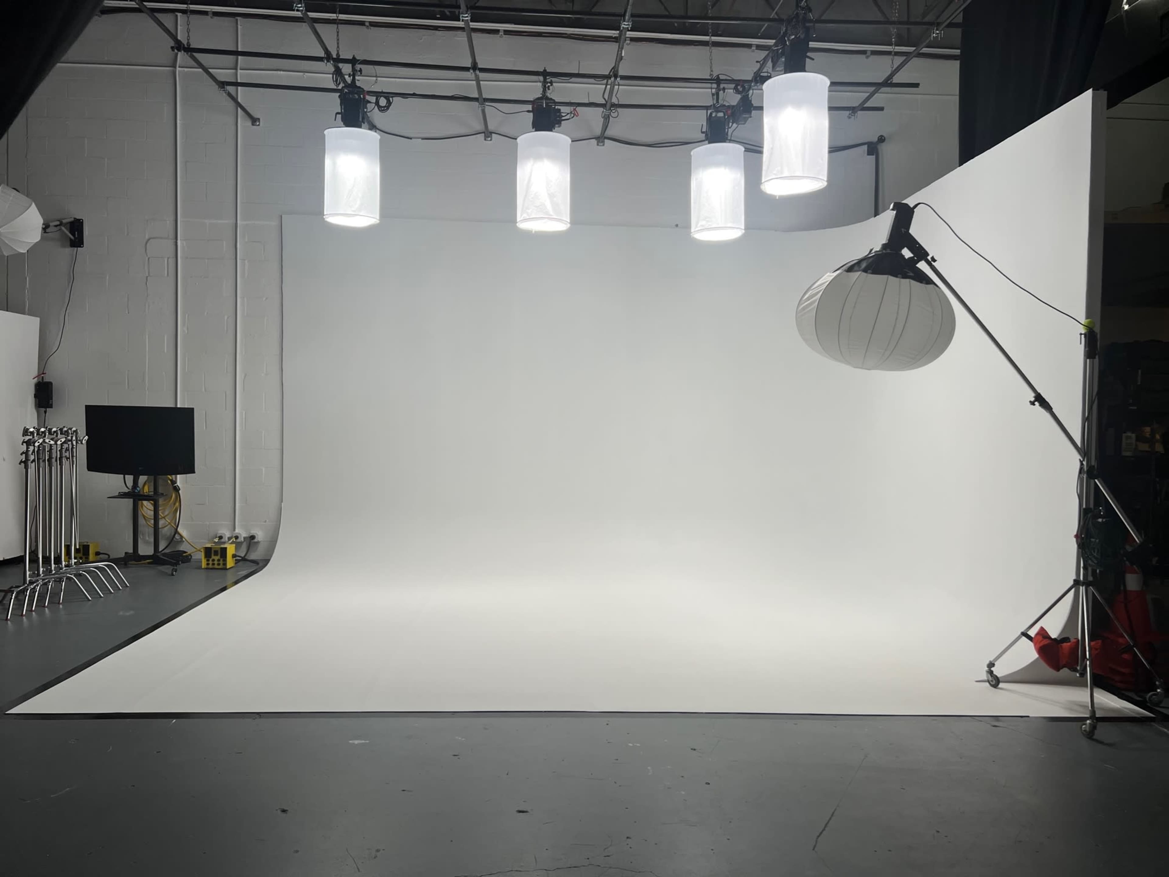 A spacious photography studio with a large white backdrop, multiple hanging lights, and a light stand with a diffuser.