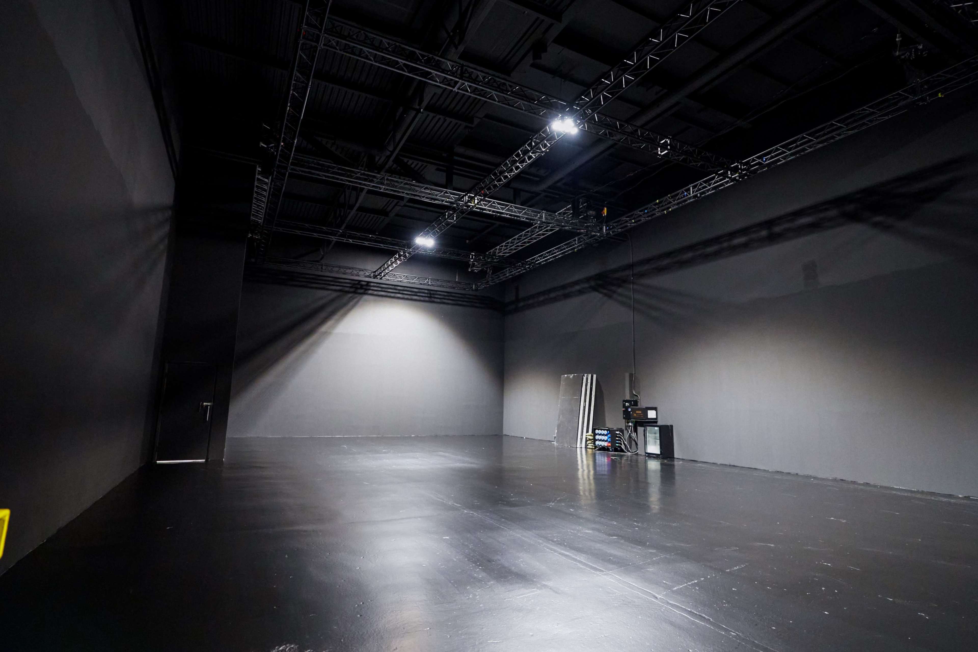 The image shows a large, empty studio space with black walls and a reflective floor, illuminated by overhead lights.