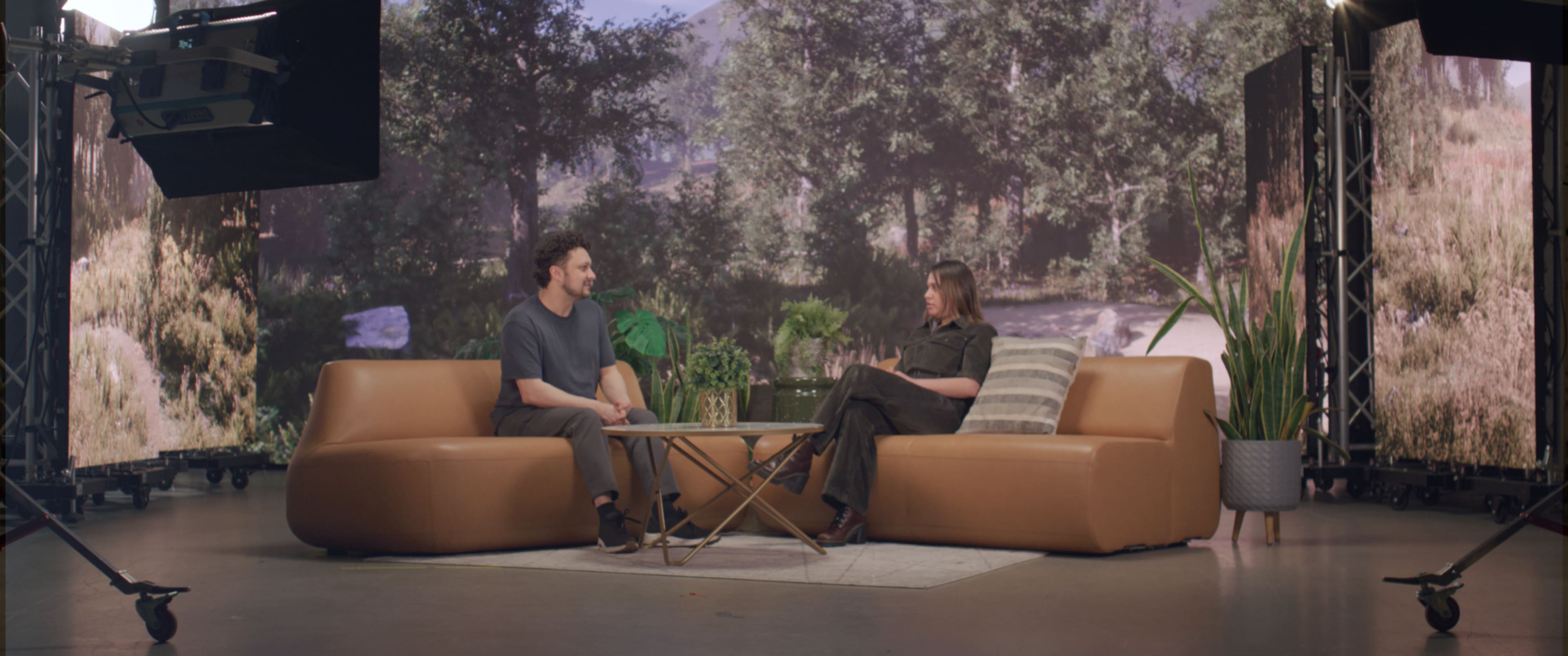 A man and a woman are seated on a brown couch in a studio setting, with a nature backdrop featuring trees and foliage behind them.