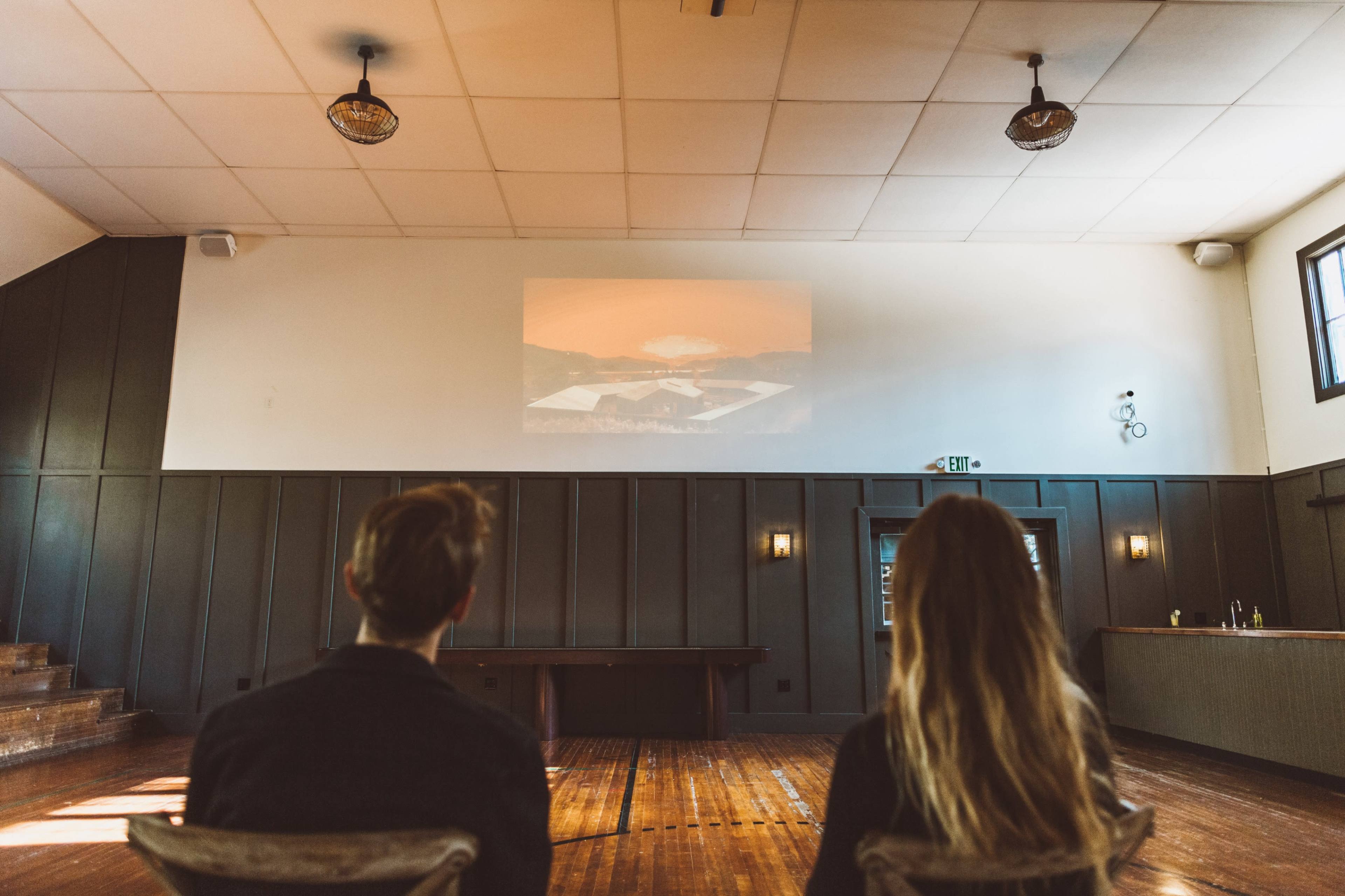 Two people sit in a room with wooden flooring, facing a projection on the wall that displays a landscape scene.