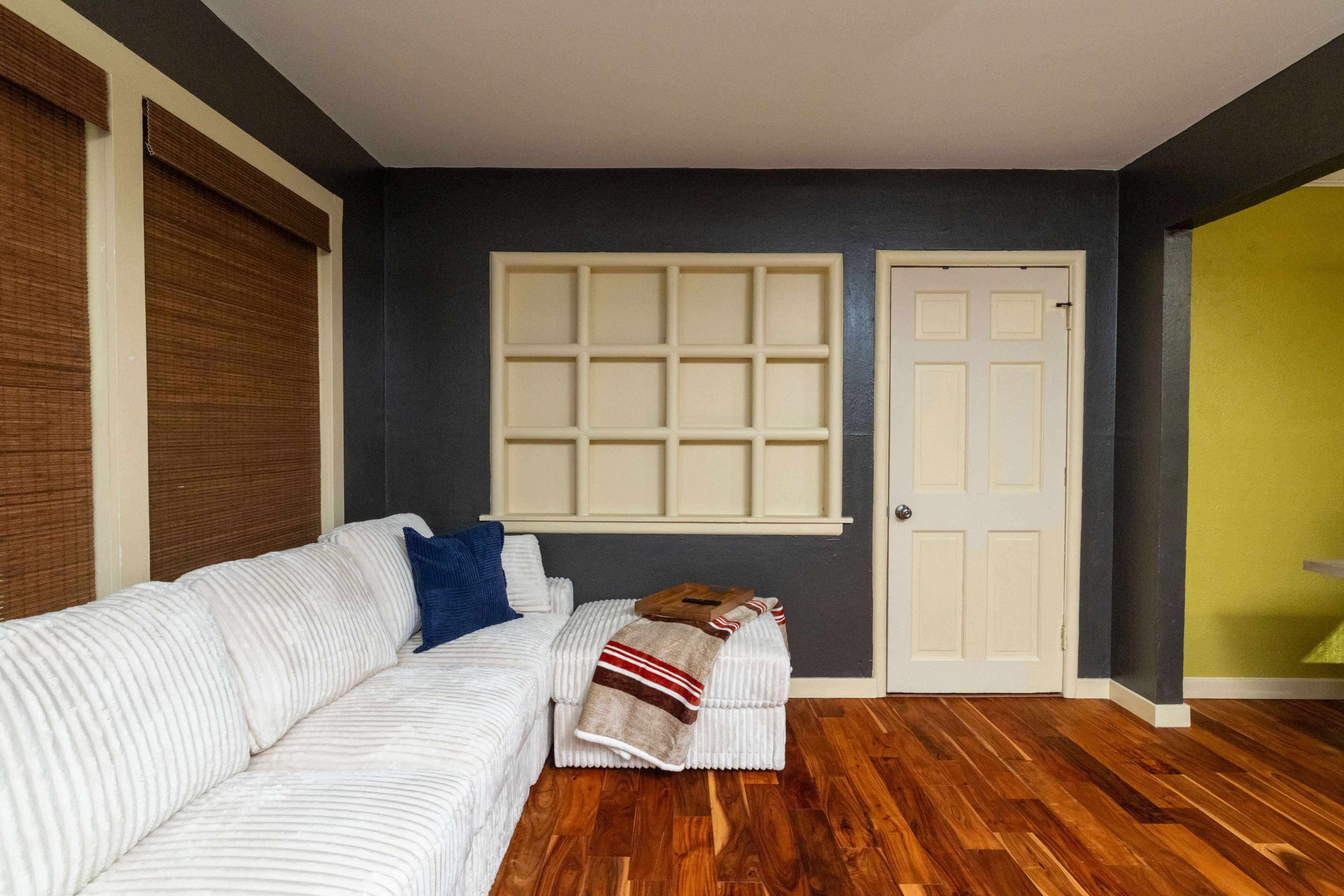 A corner of a room features a white sectional sofa, a decorative wall shelf, a closed door, and hardwood flooring.